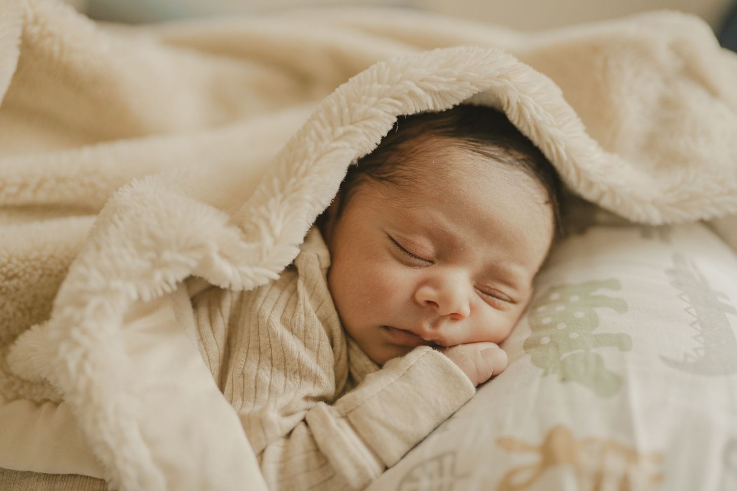 Newborn baby sleeping peacefully wrapped in a soft cream blanket with hands tucked under chin.