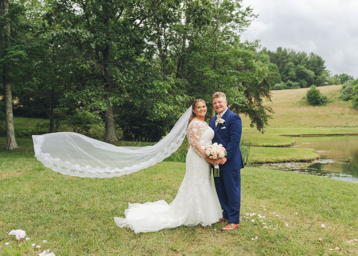Bride and groom pose outdoors on green grass, her veil flowing in the wind, with trees and a pond backdrop.