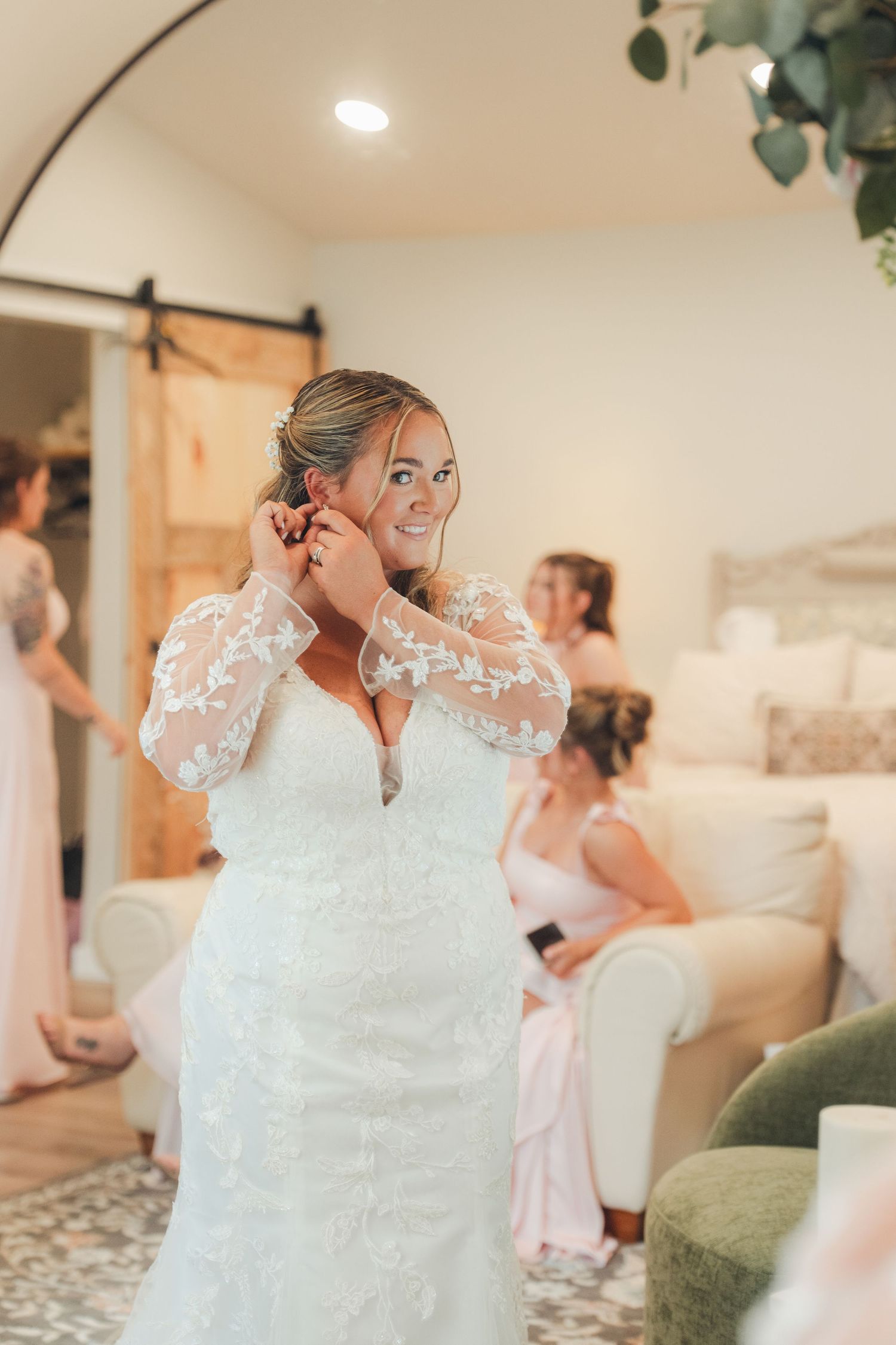 Bride in lace wedding gown adjusting earring while bridesmaids in pink dresses relax in elegant bridal suite.