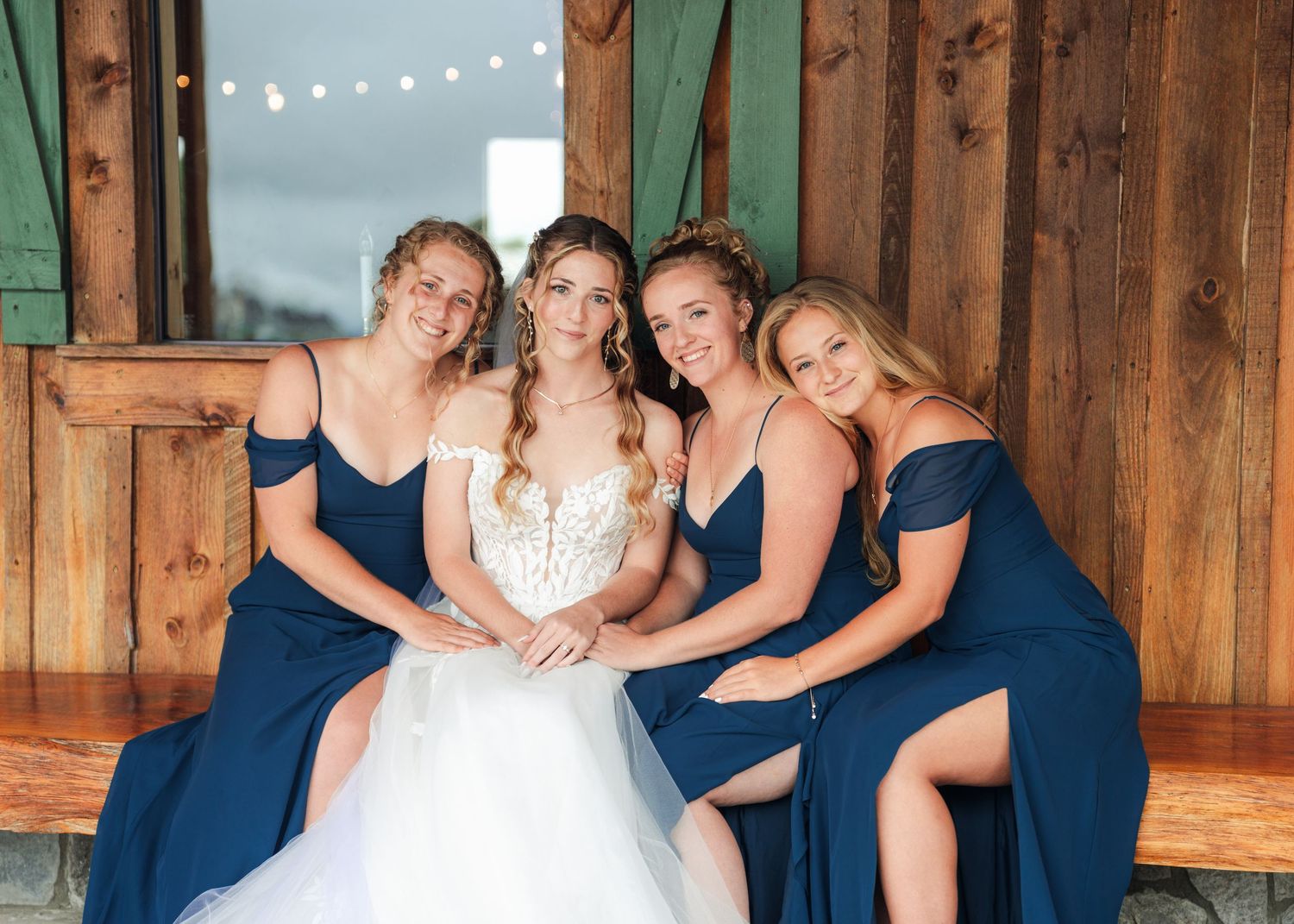 Bride in white gown poses with three bridesmaids in navy blue off-shoulder dresses inside a rustic barn venue.