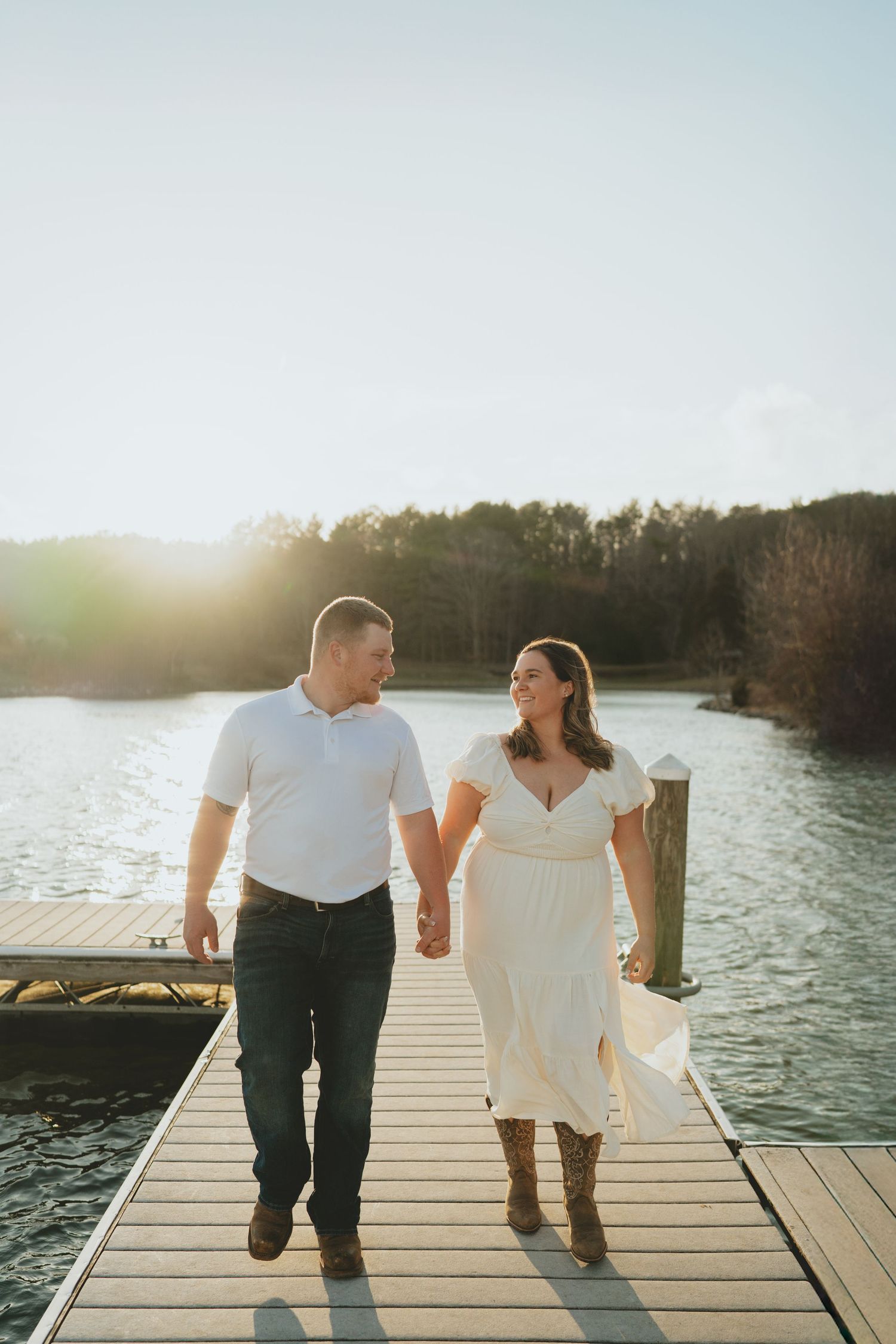 Happy couple walking hand-in-hand on a lakeside dock smiling at each other during golden hour.