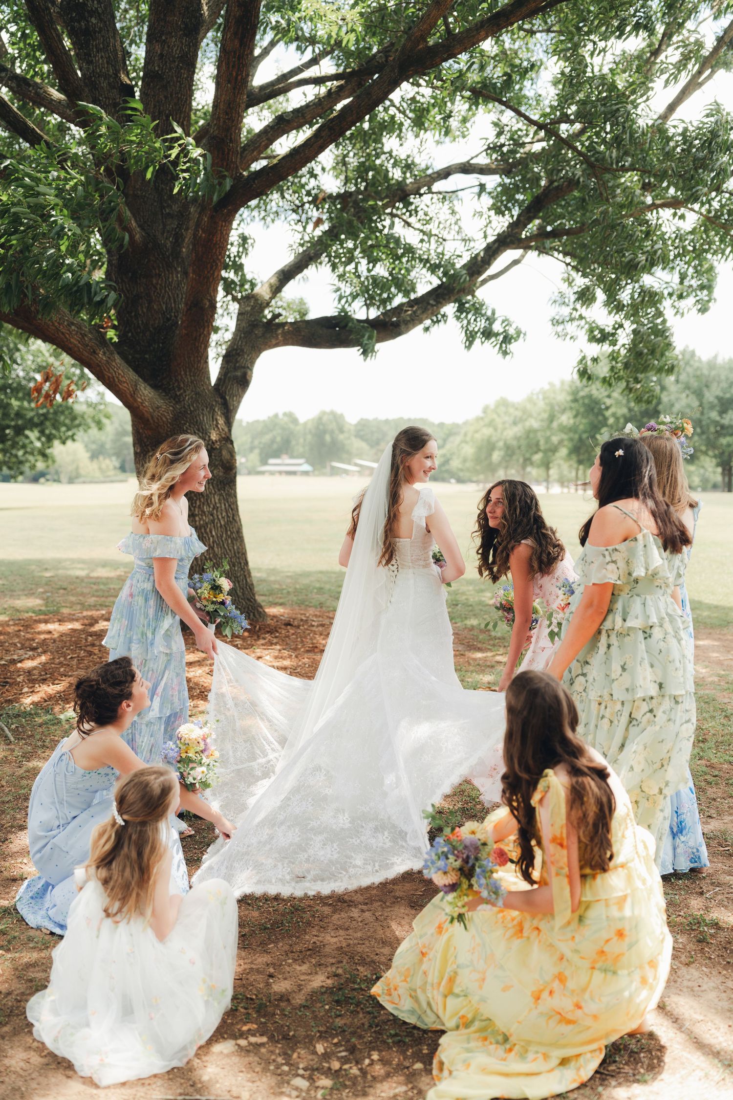 Bride in white gown surrounded by bridesmaids in floral dresses under a large tree at an outdoor wedding.