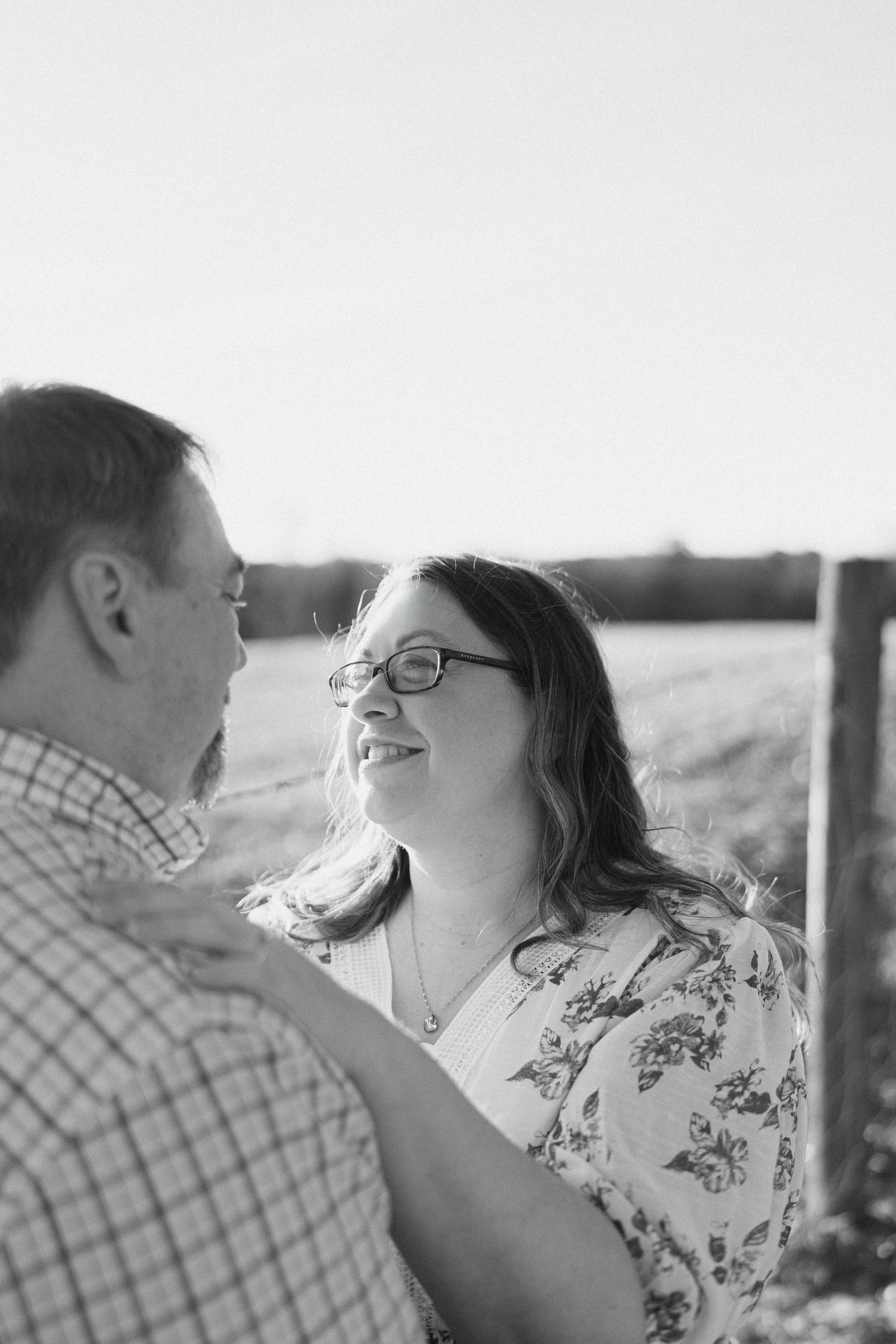 Black and white photo of a smiling couple gazing at each other near a wooden fence post.