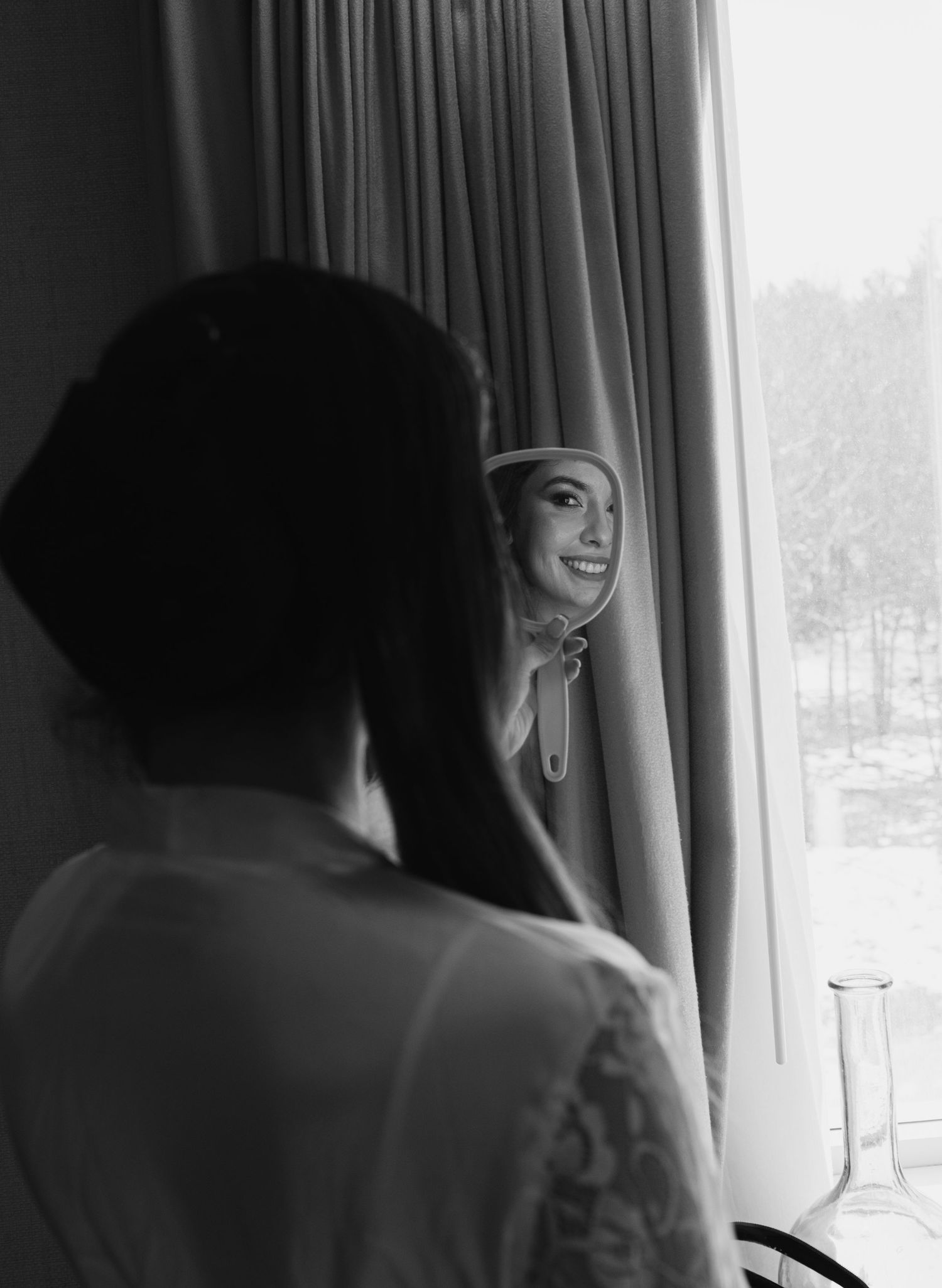 Black and white photo of a smiling bride checking her appearance in a handheld mirror.