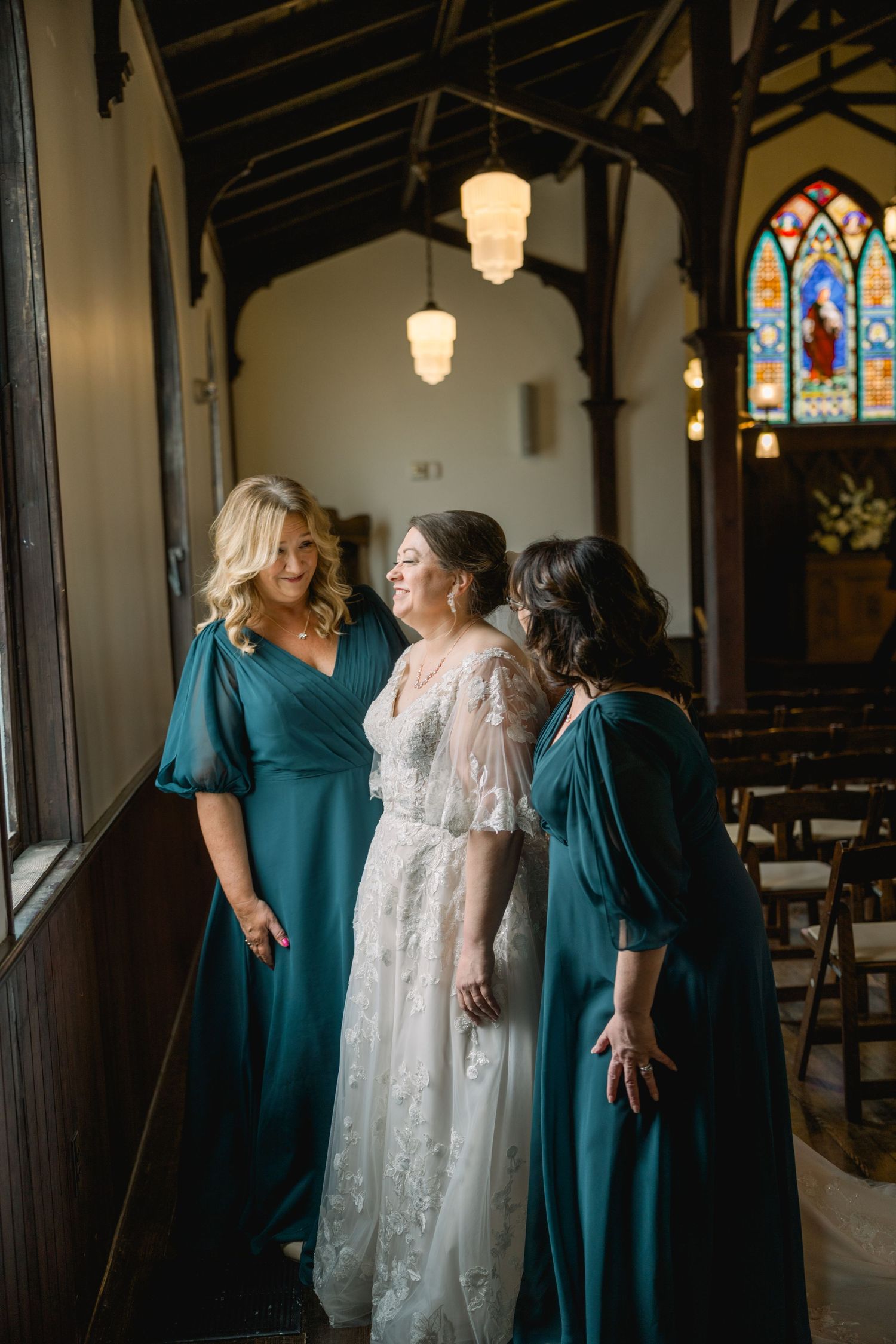 Bride in lace gown laughing with bridesmaids in teal dresses inside historic church.