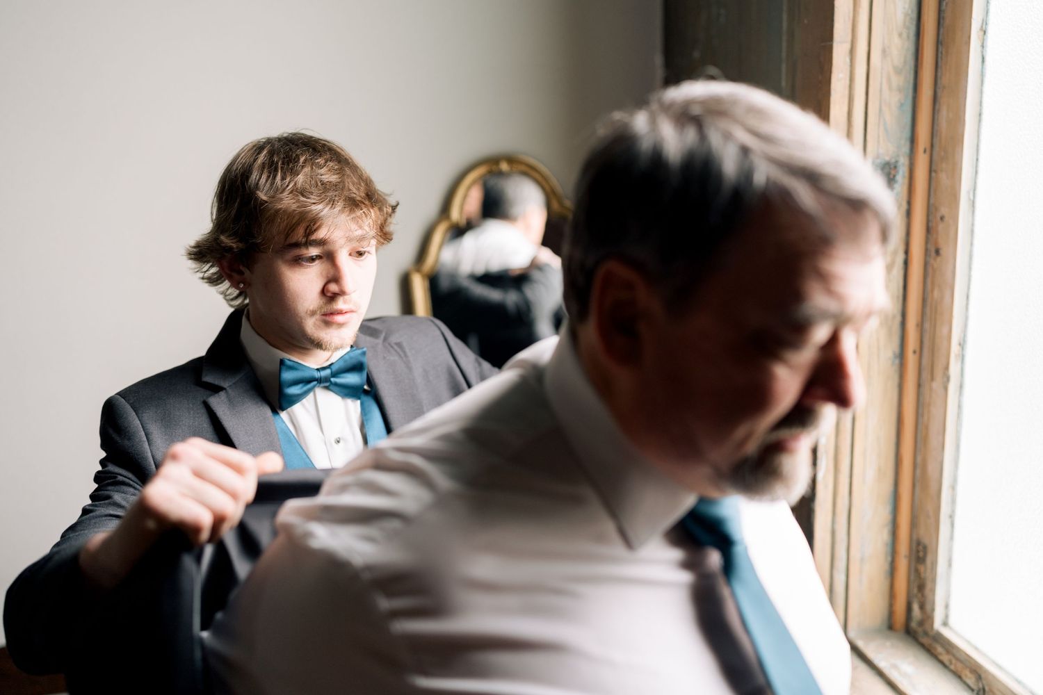 Young groomsman in teal bow tie adjusting his jacket while an older man stands near a window at wedding.