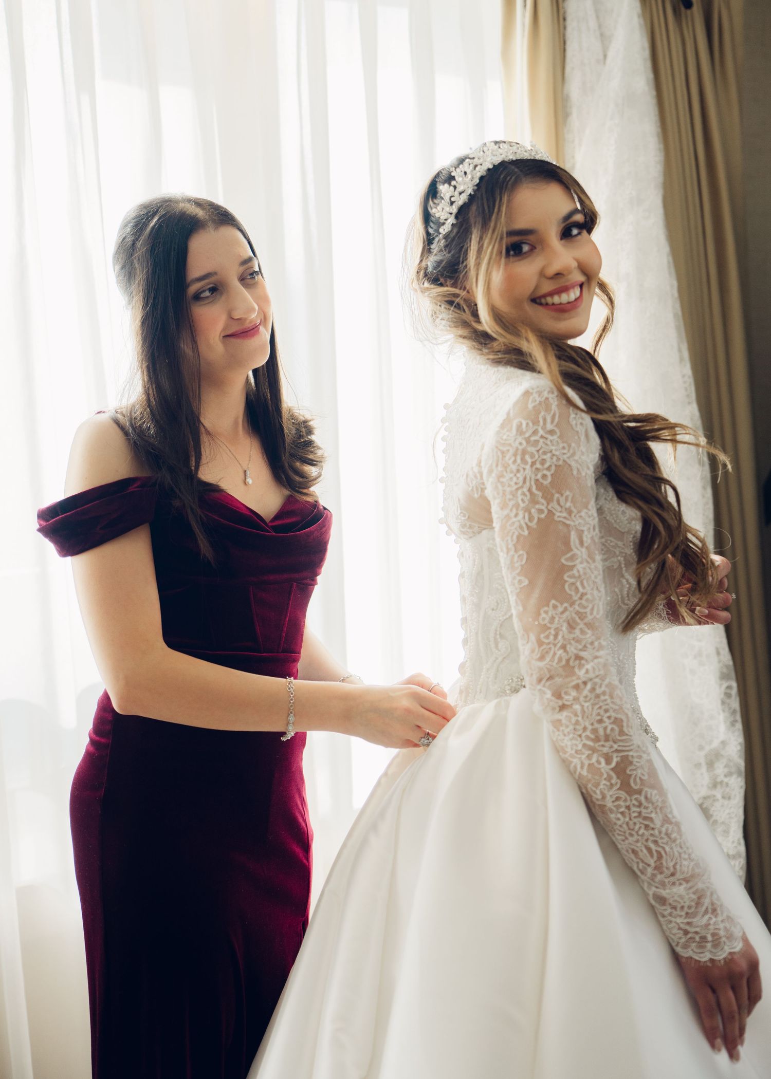Bridesmaid in burgundy velvet dress helping smiling bride in lace wedding gown get dressed.