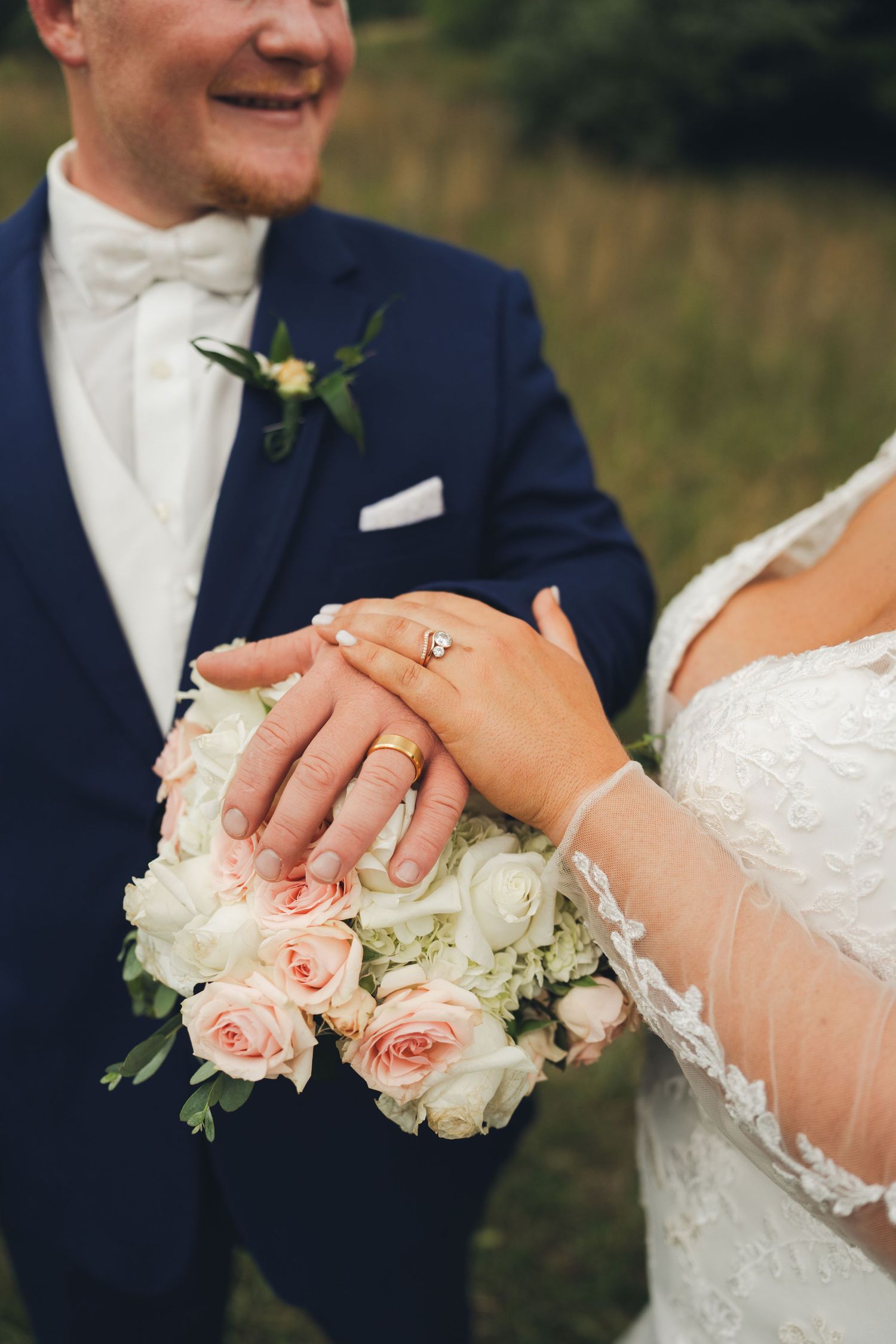 Bride and groom display wedding rings resting hands on white and pink rose bouquet, groom smiling in navy suit.
