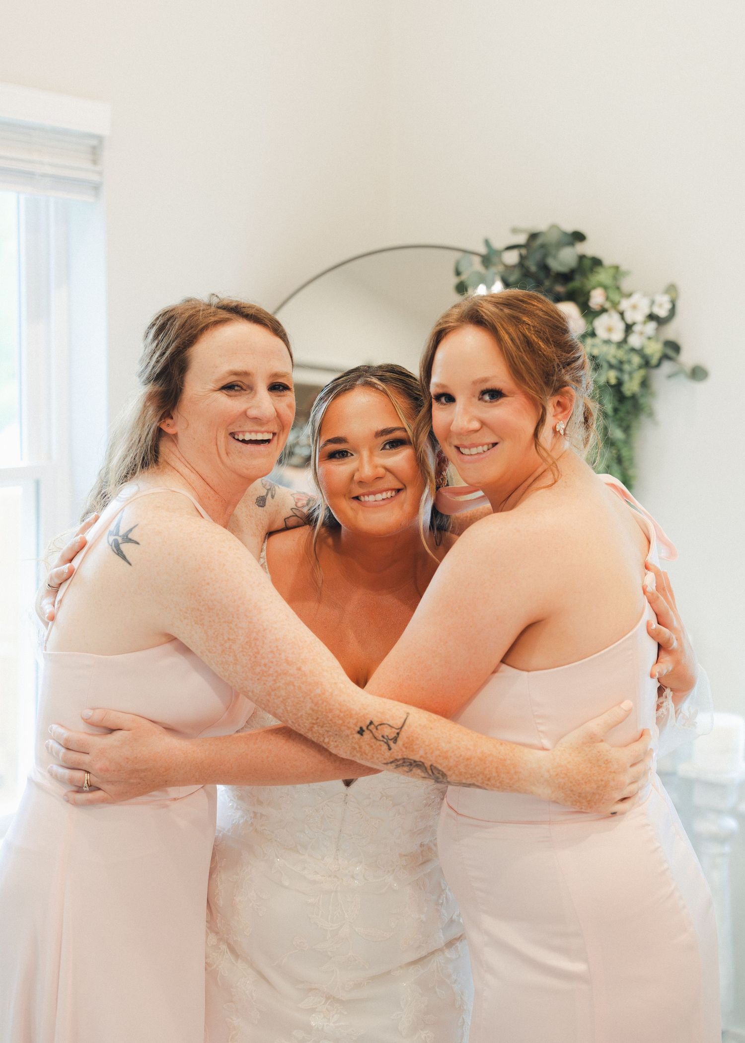 Bride in white gown hugged by two bridesmaids in blush pink dresses, smiling together on wedding day.