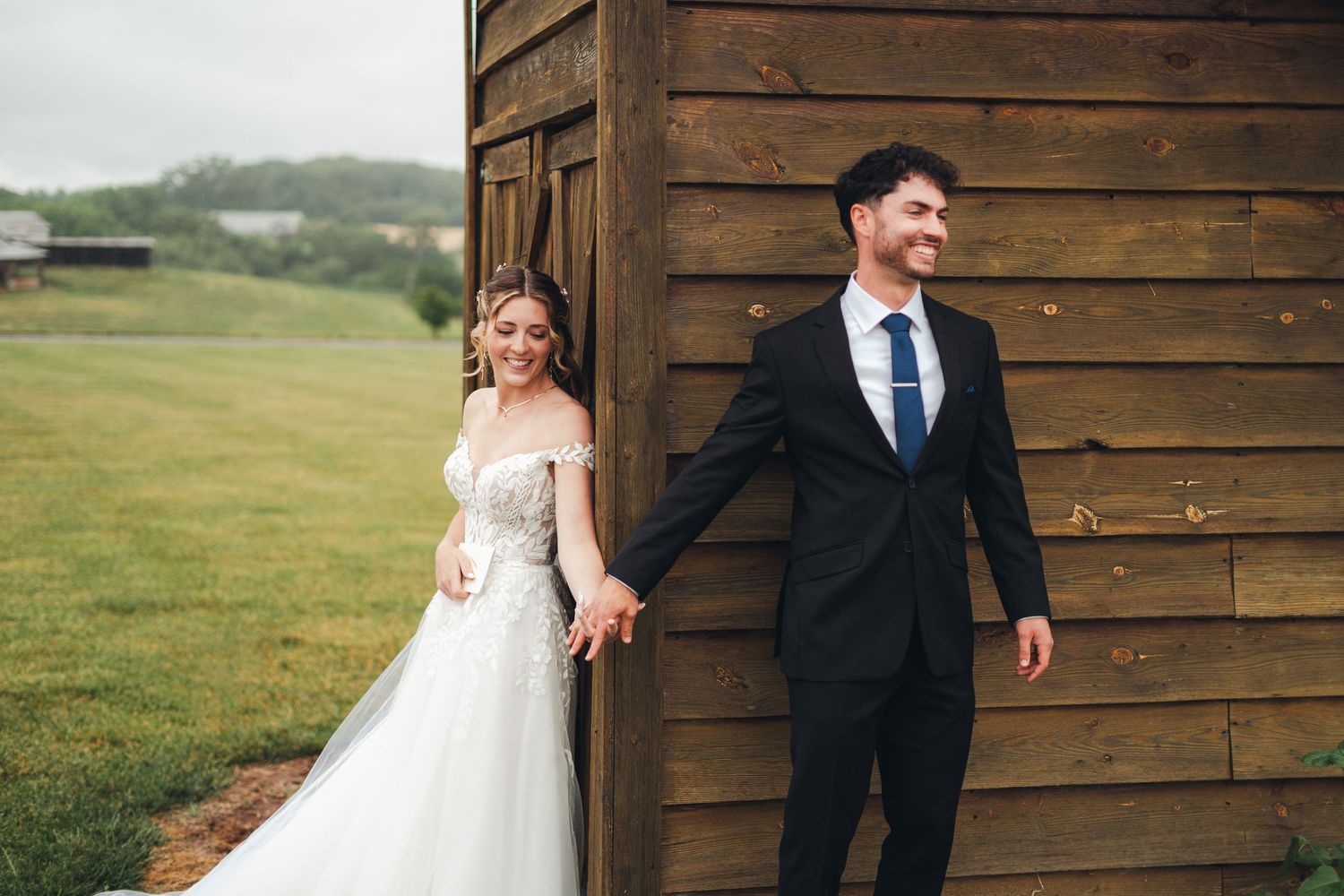 Bride in white lace gown holds groom's hand in black suit by rustic wooden barn on green lawn.