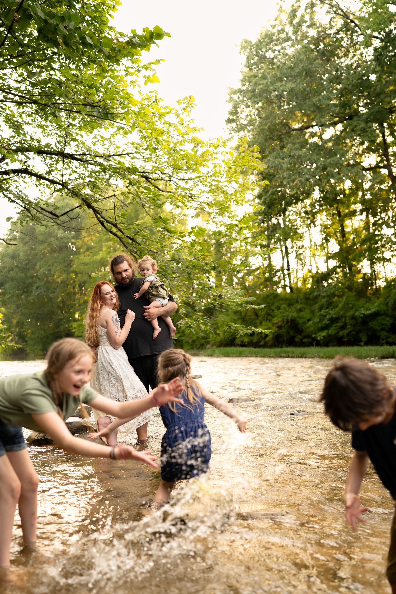 Family playing and splashing in a shallow river surrounded by lush green trees on a sunny day.