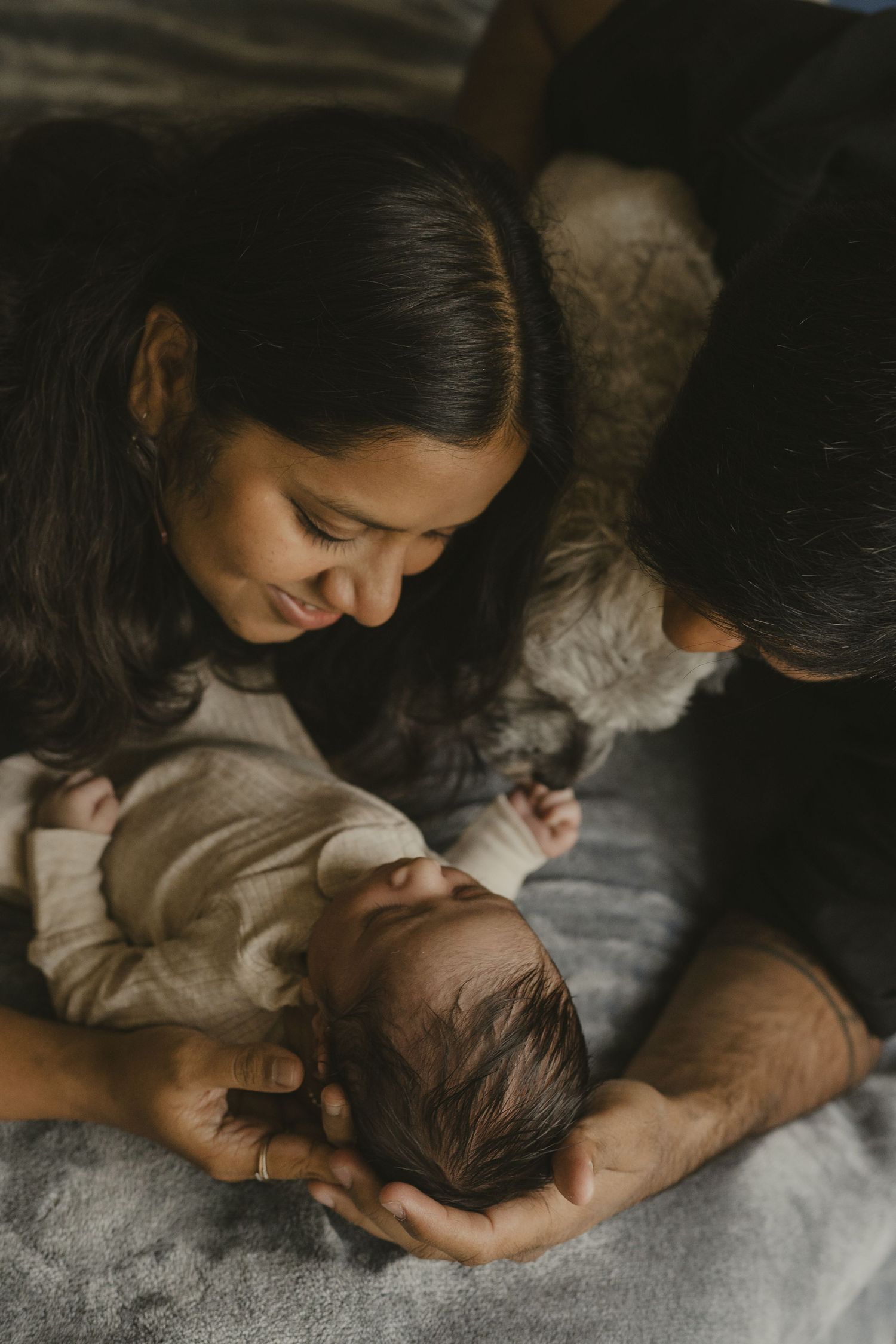 Parents lovingly gazing down at sleeping newborn baby with family dog nearby in dim light.