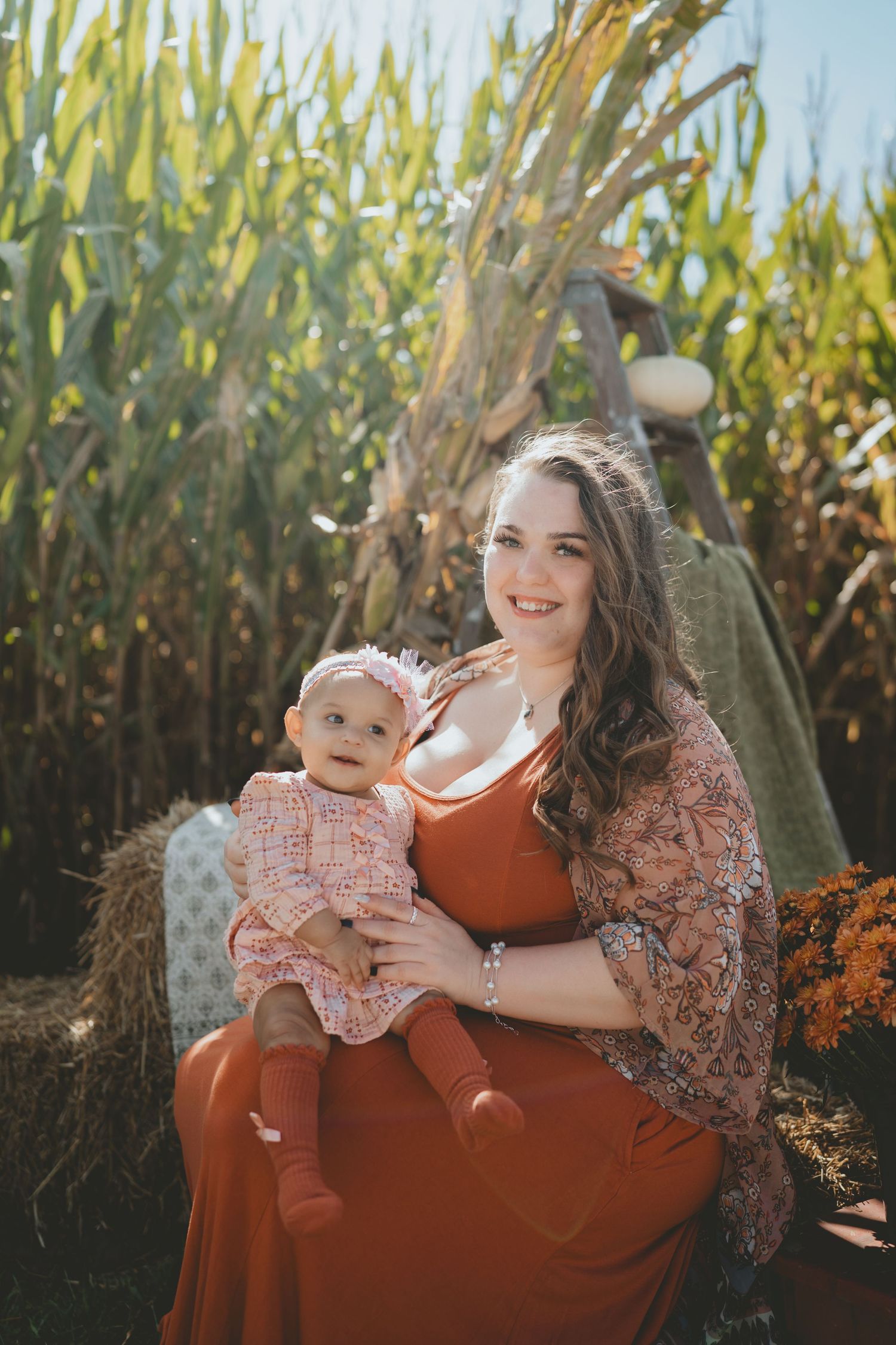 Smiling mother in rust orange dress holding baby girl on a hay bale beside a sunny cornfield.