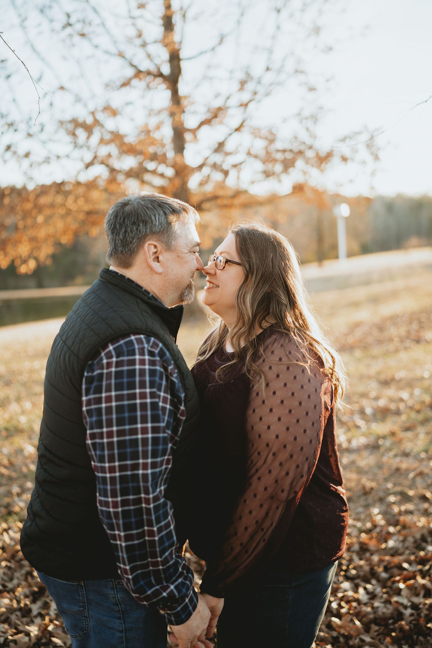 Couple leaning in for a kiss while holding hands among autumn leaves in golden sunlight.