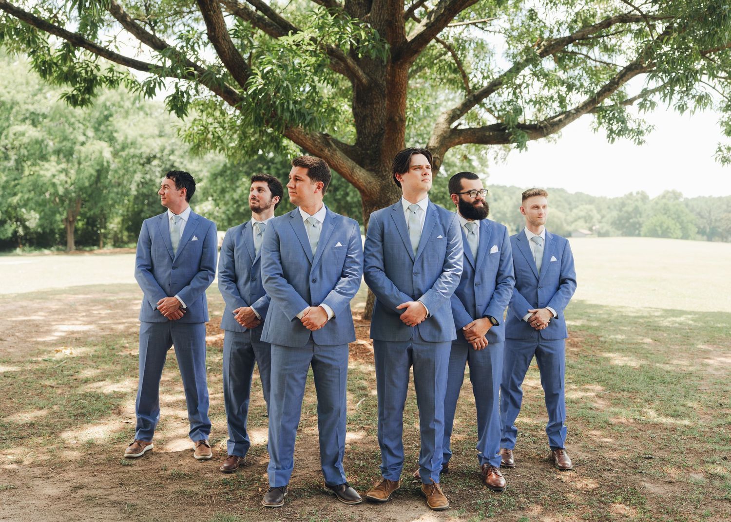 Groom and groomsmen in matching blue suits standing outdoors under large tree at wedding ceremony.
