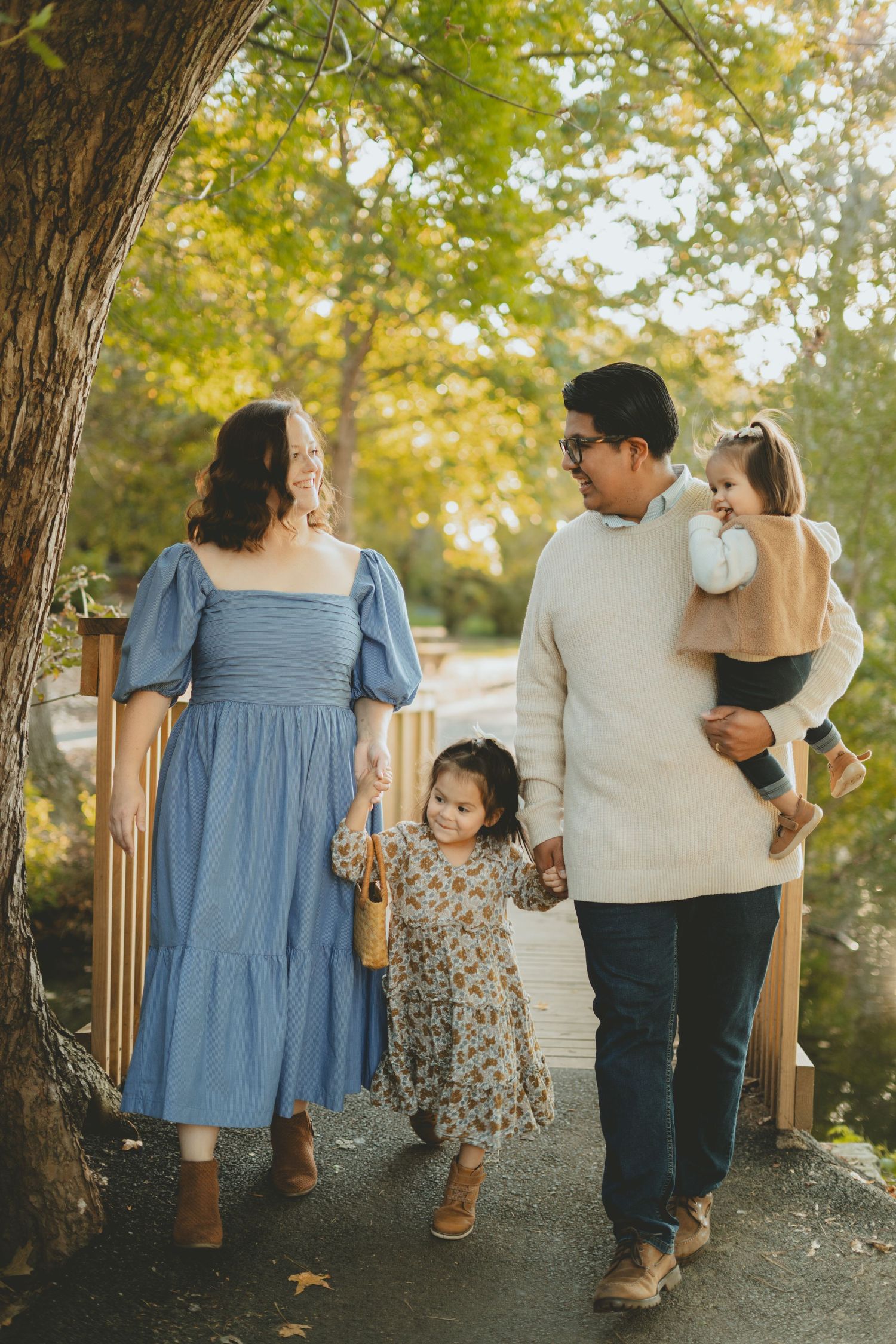 Happy family of four walking hand in hand along a sunlit autumn path near a wooden bridge.