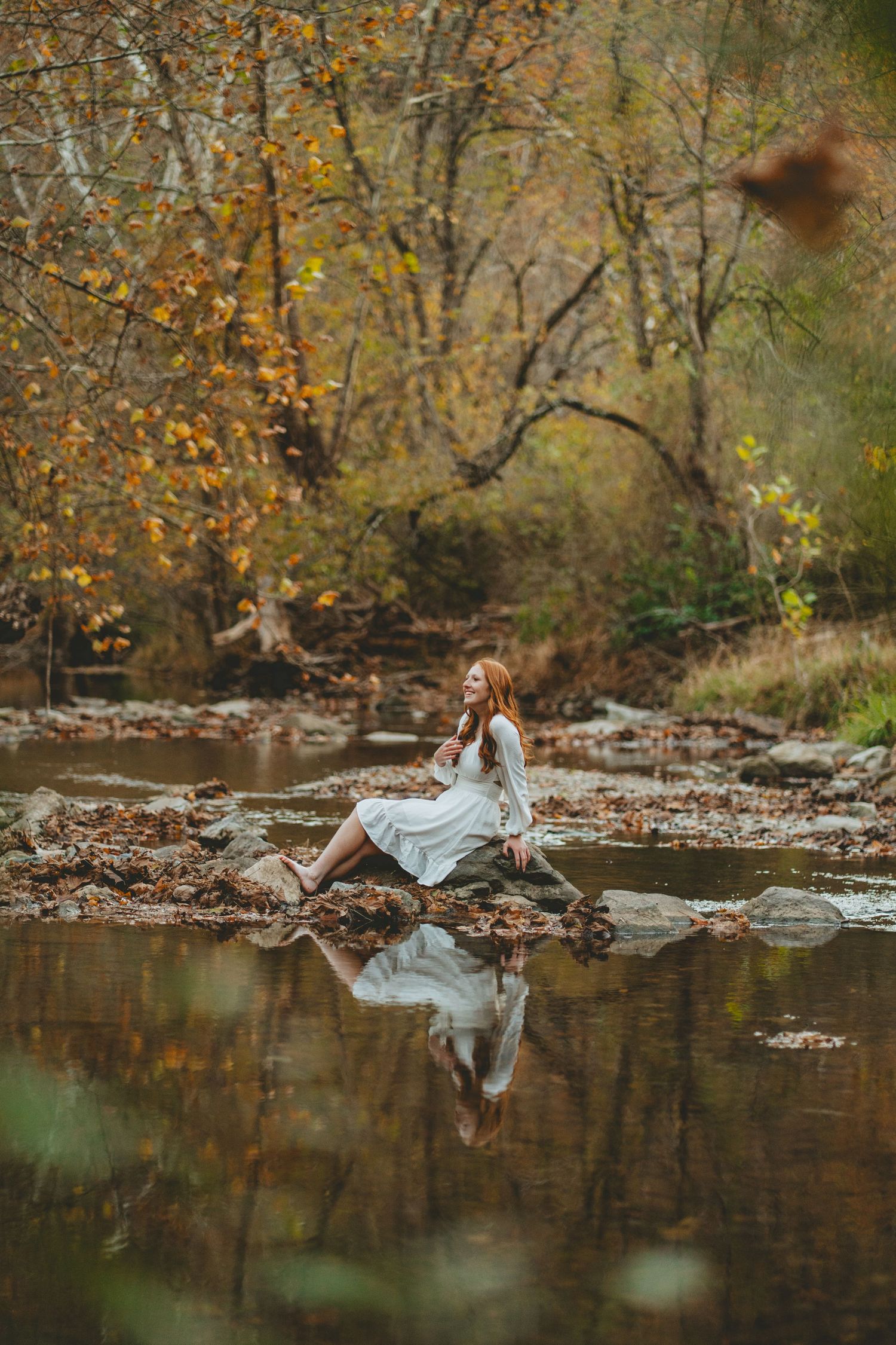 Young woman in white dress sitting on rocks beside a tranquil autumn forest creek with her reflection in the water.