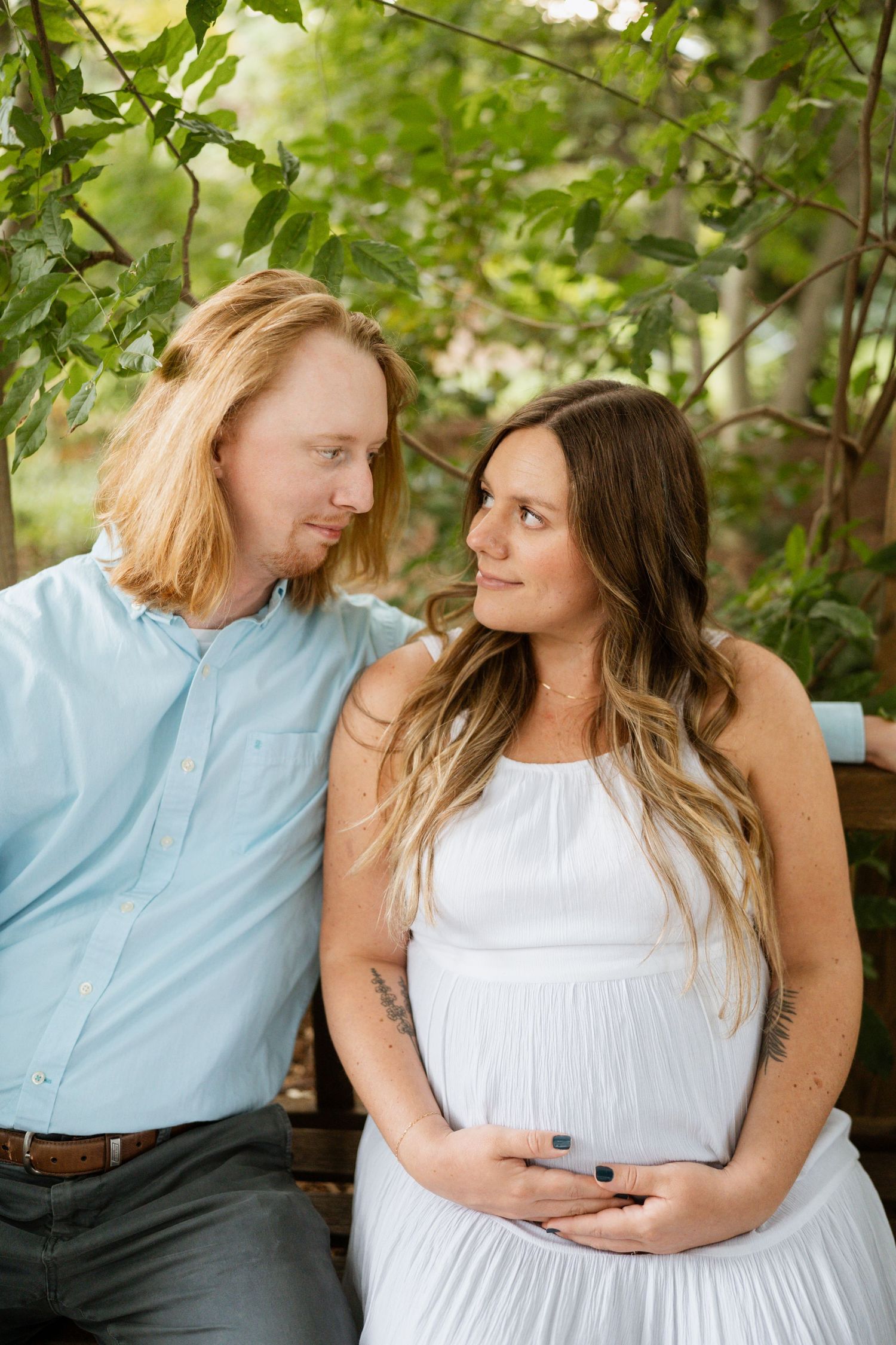 Pregnant woman and partner sitting on bench surrounded by green foliage.