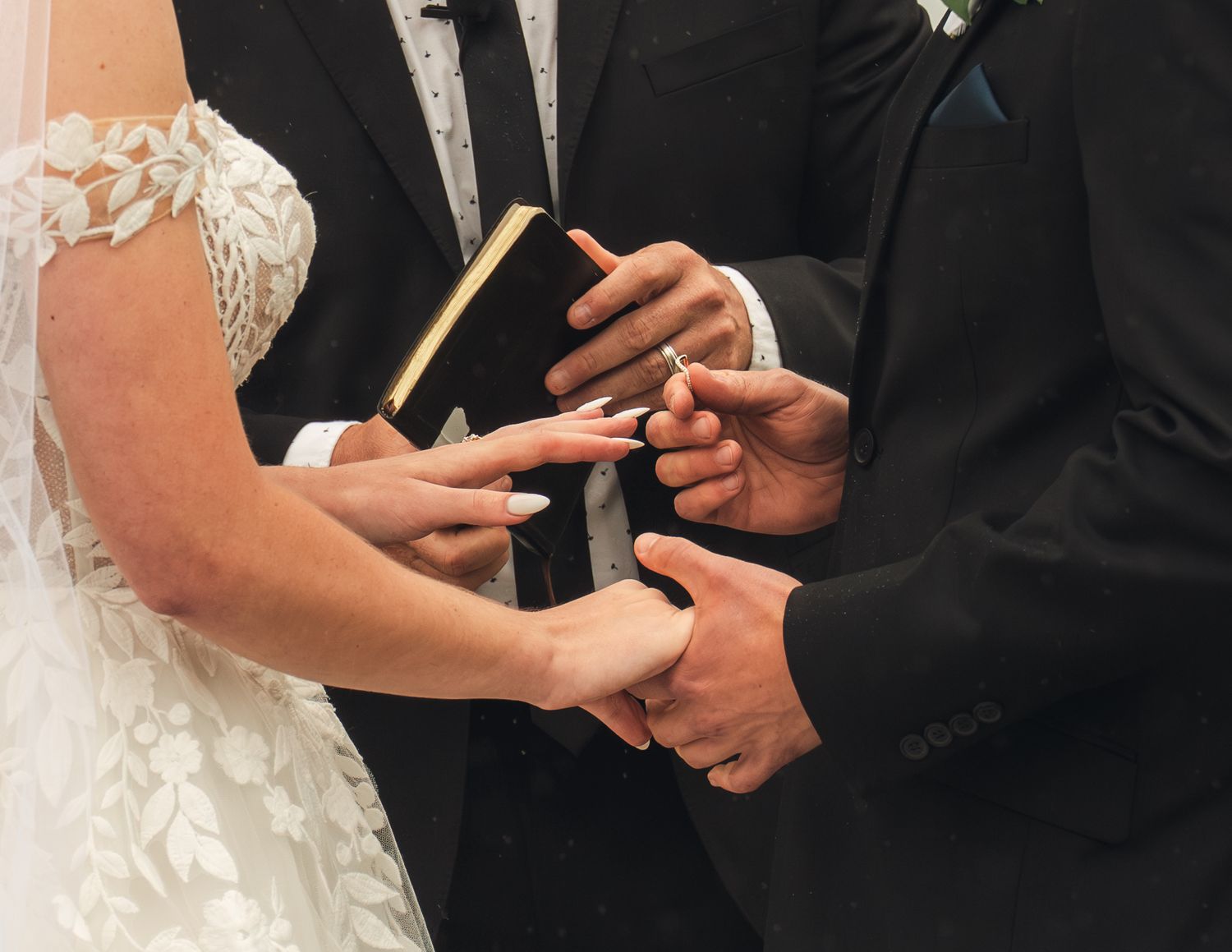 Groom placing a wedding ring on brides finger during ceremony, officiant holding Bible, elegant white lace dress.