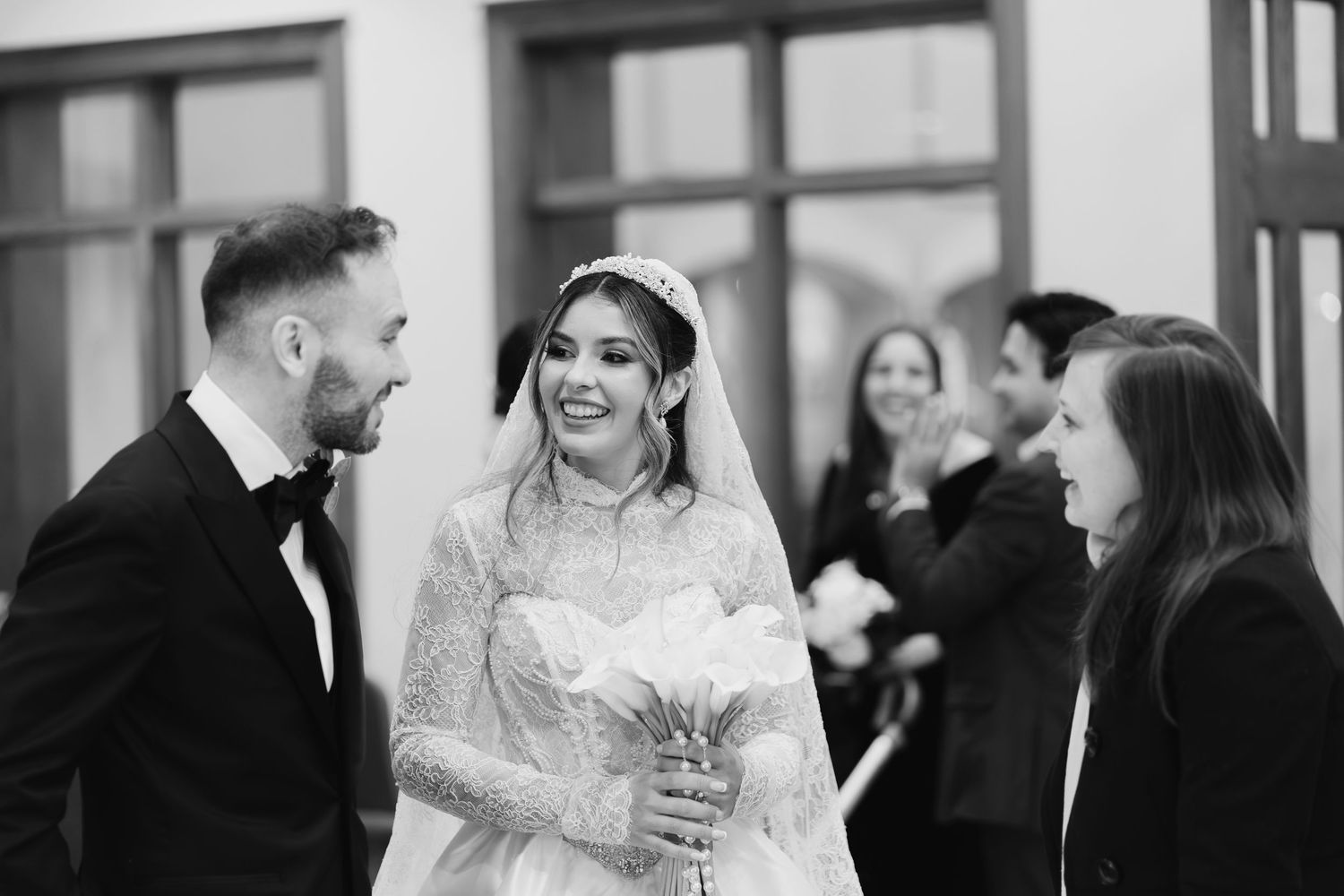 Smiling bride in lace gown holds bouquet while greeting guests after church ceremony.