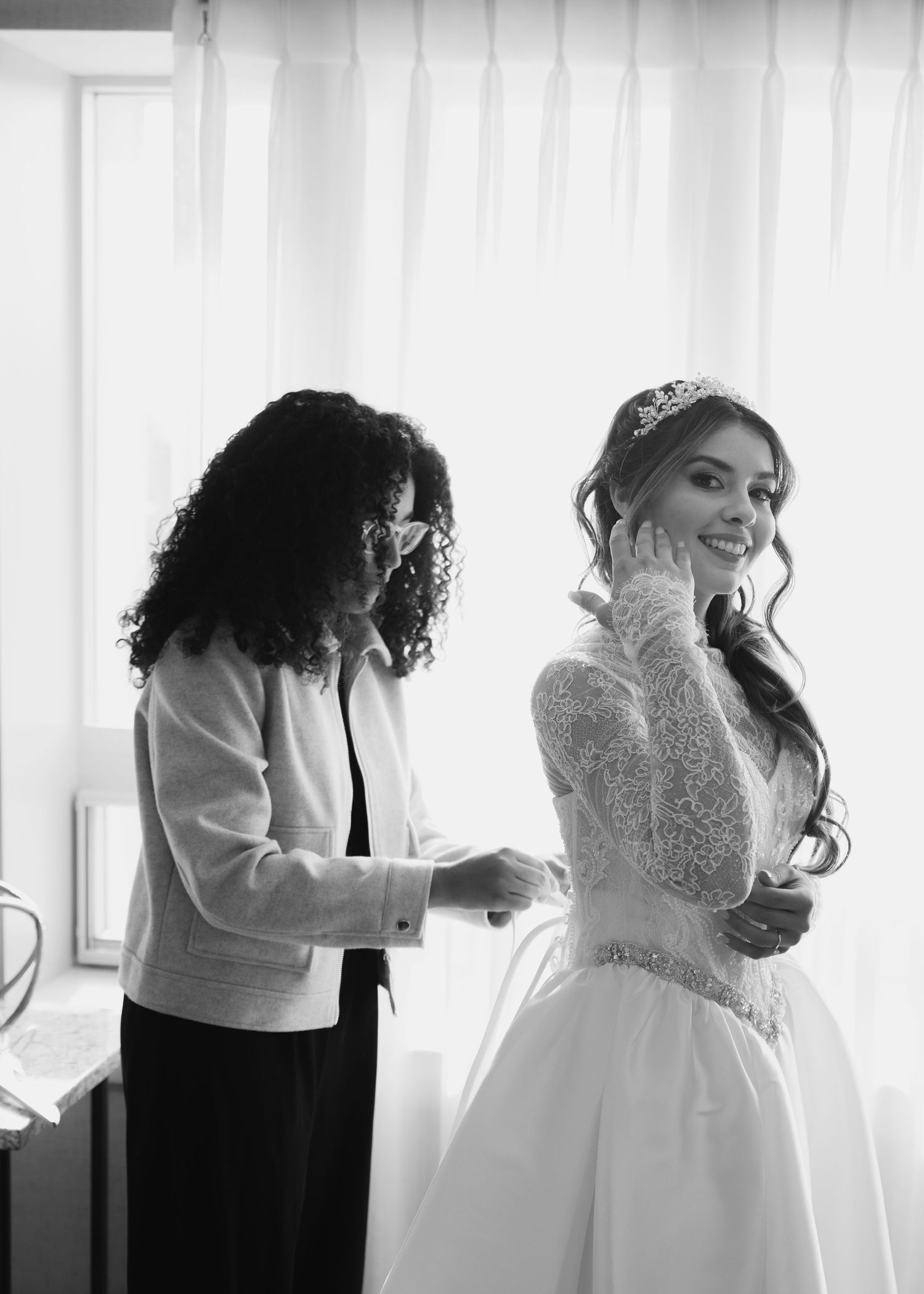 Black and white photo of a bride smiling as her mother fastens her lace wedding gown.