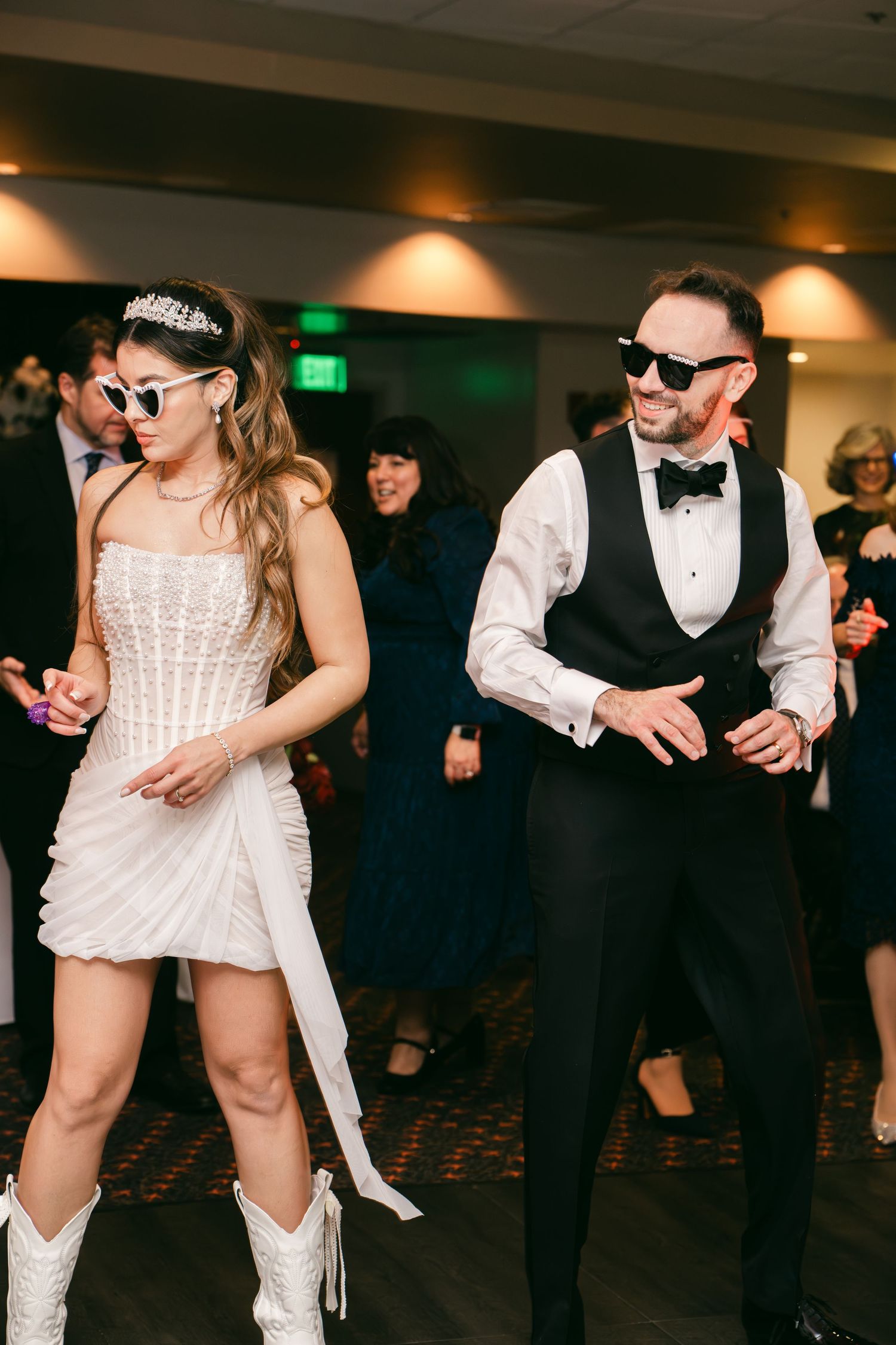 Bride in short white dress and groom in tuxedo vest dance wearing sunglasses at reception.
