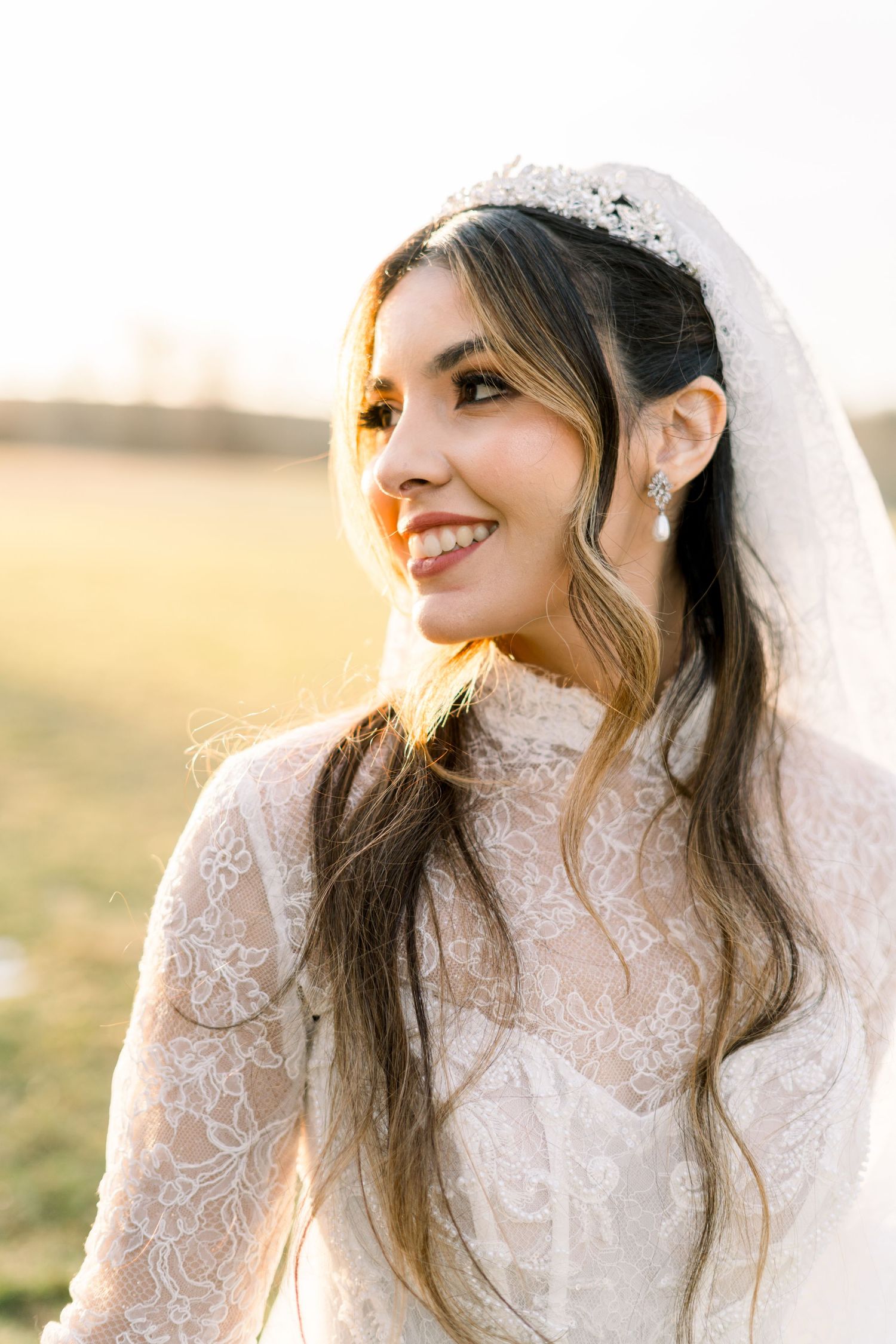 Radiant bride in lace wedding gown and veil smiling softly in warm golden sunset light outdoors.