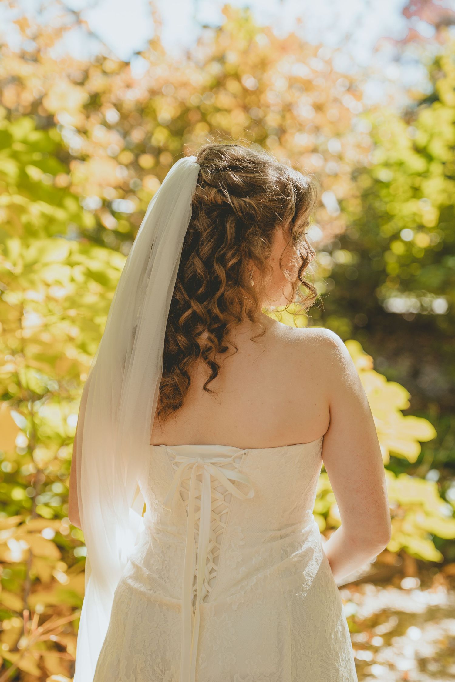 Bride seen from behind in a lace corset wedding gown with a veil amid vibrant autumn foliage.