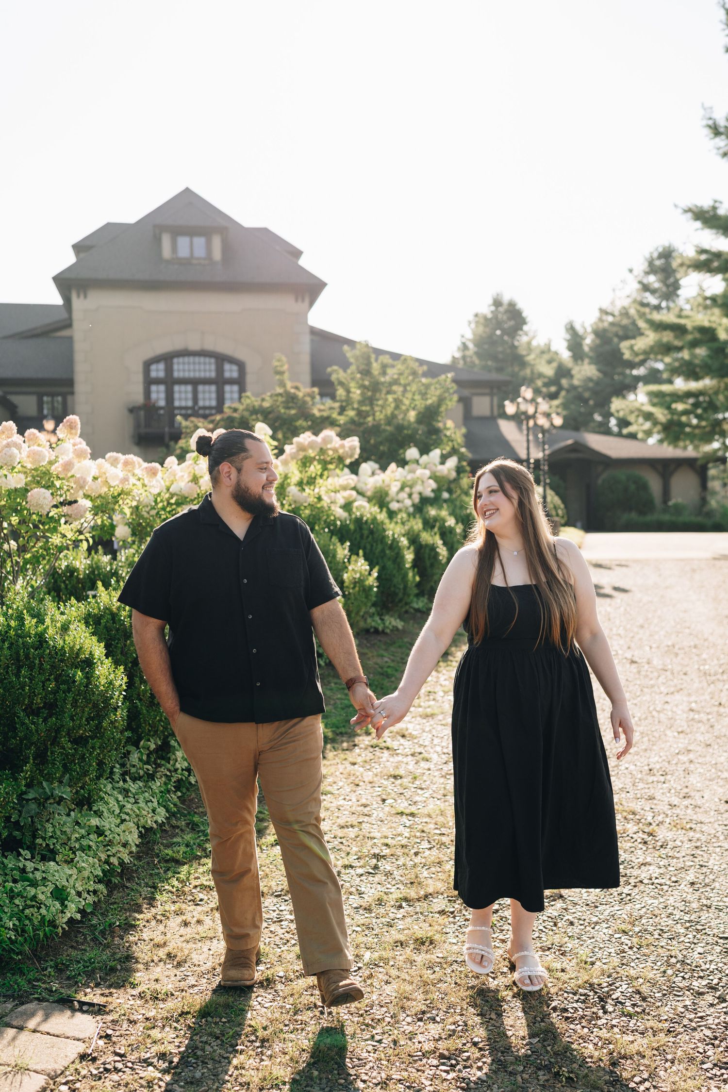 Engaged couple holding hands and walking past blooming hydrangeas in front of an elegant building.