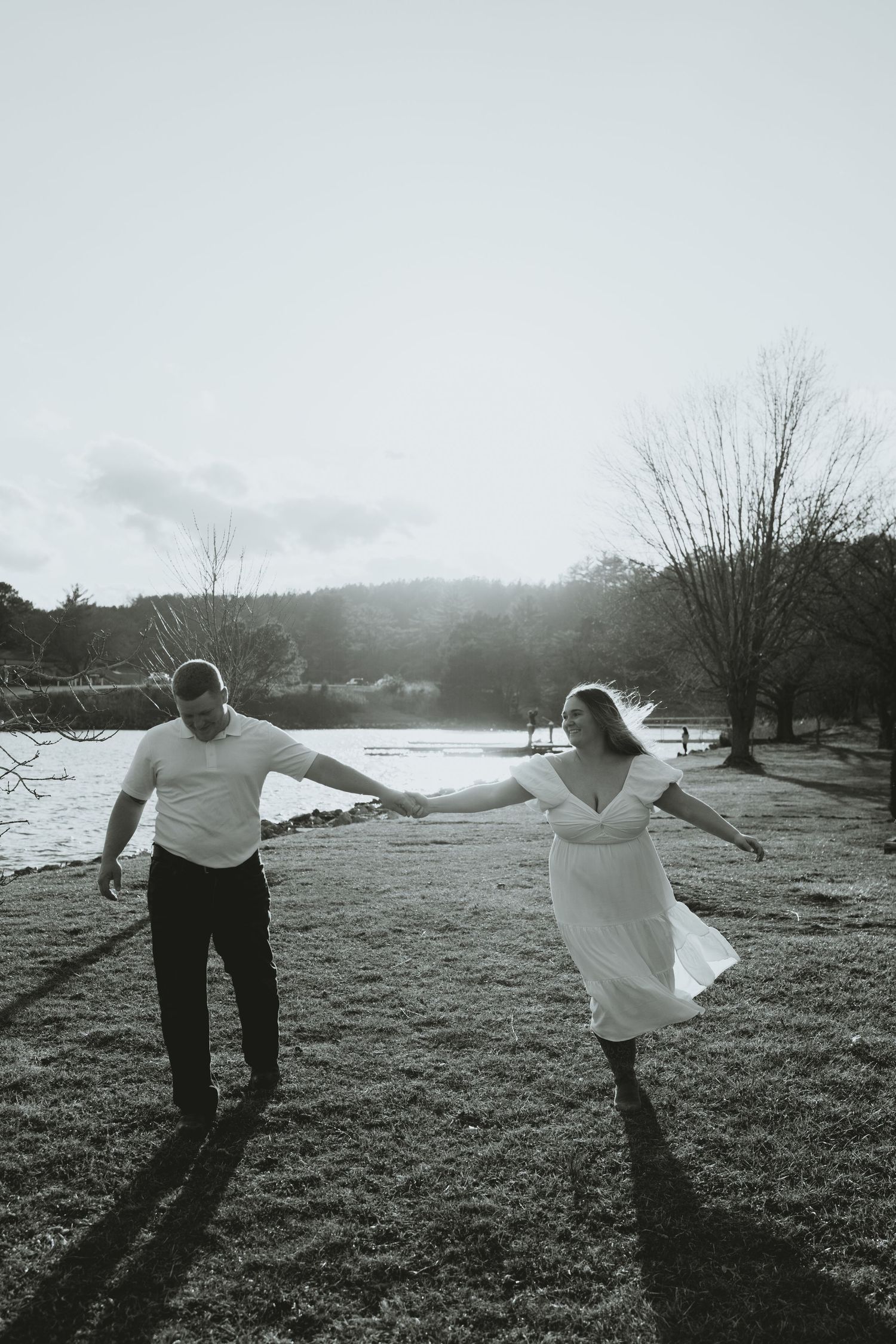 Black and white photo of couple playfully running through a field near a lake holding hands.