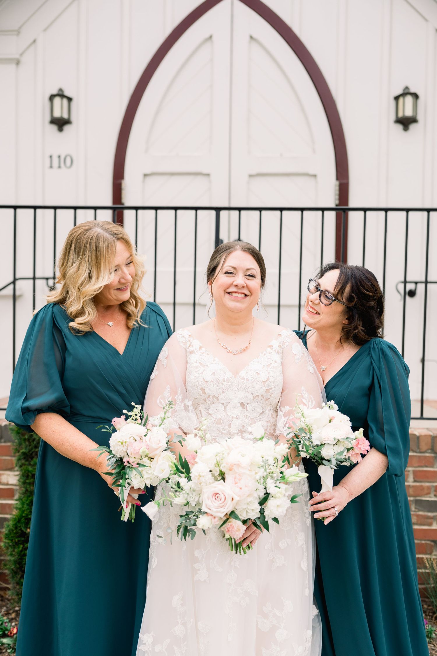 Bride in lace gown with two bridesmaids in teal dresses outside white church.