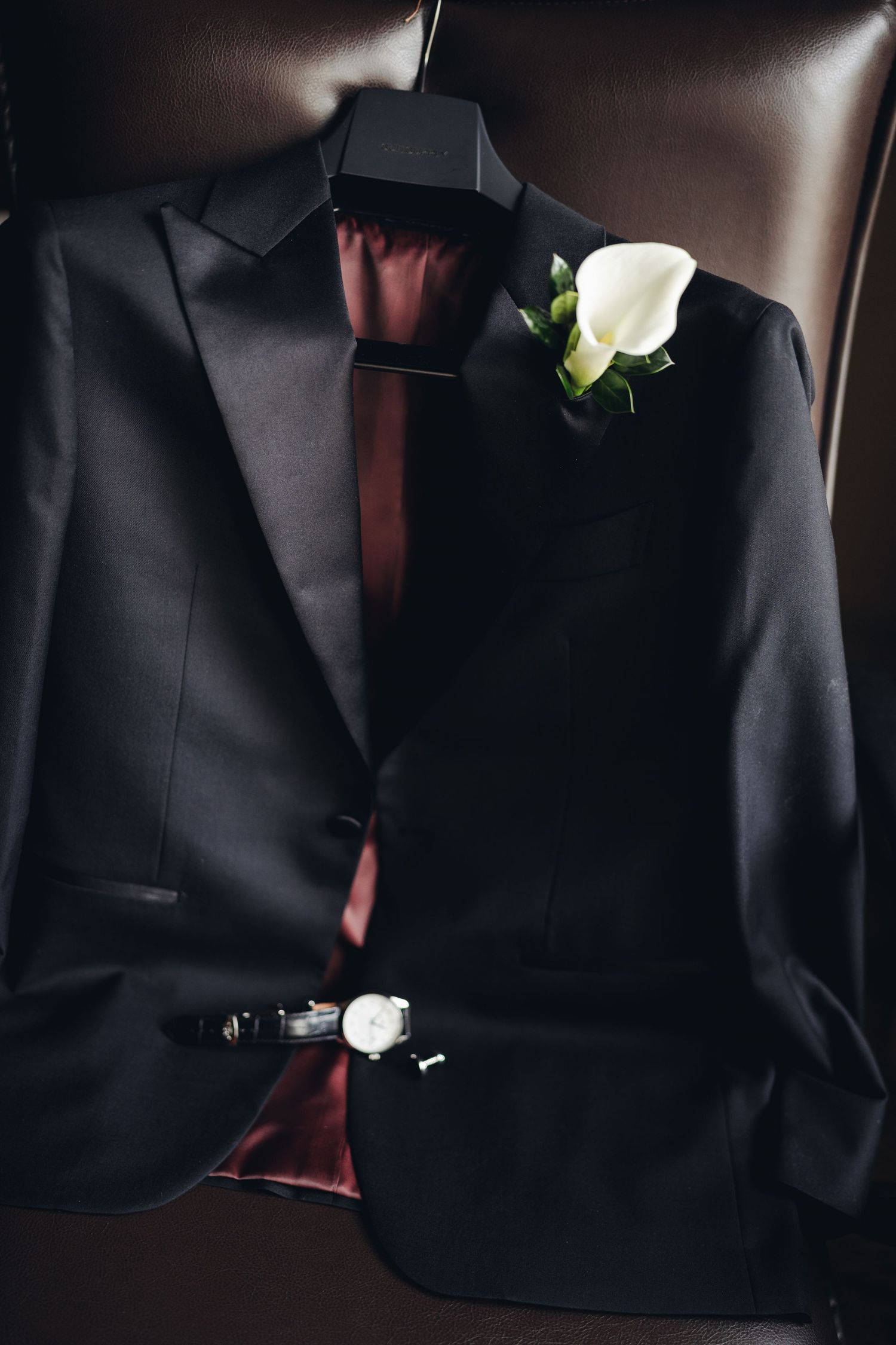 Black tuxedo jacket with white calla lily boutonniere, watch, and cufflinks displayed on leather chair.