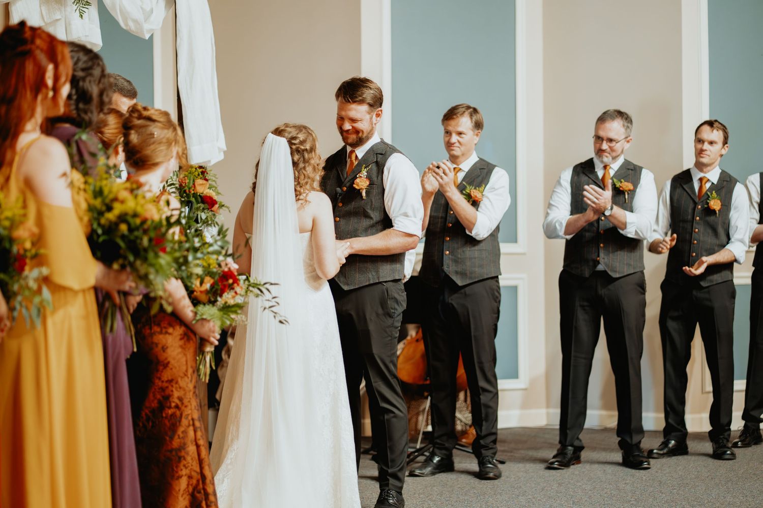 Groom laughs joyfully with groomsmen applauding during a lighthearted wedding ceremony moment.