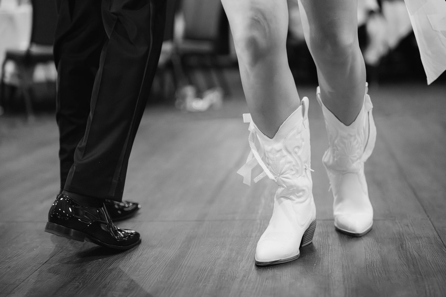 Close-up of bride in white cowboy boots and groom in dress shoes dancing at wedding reception.