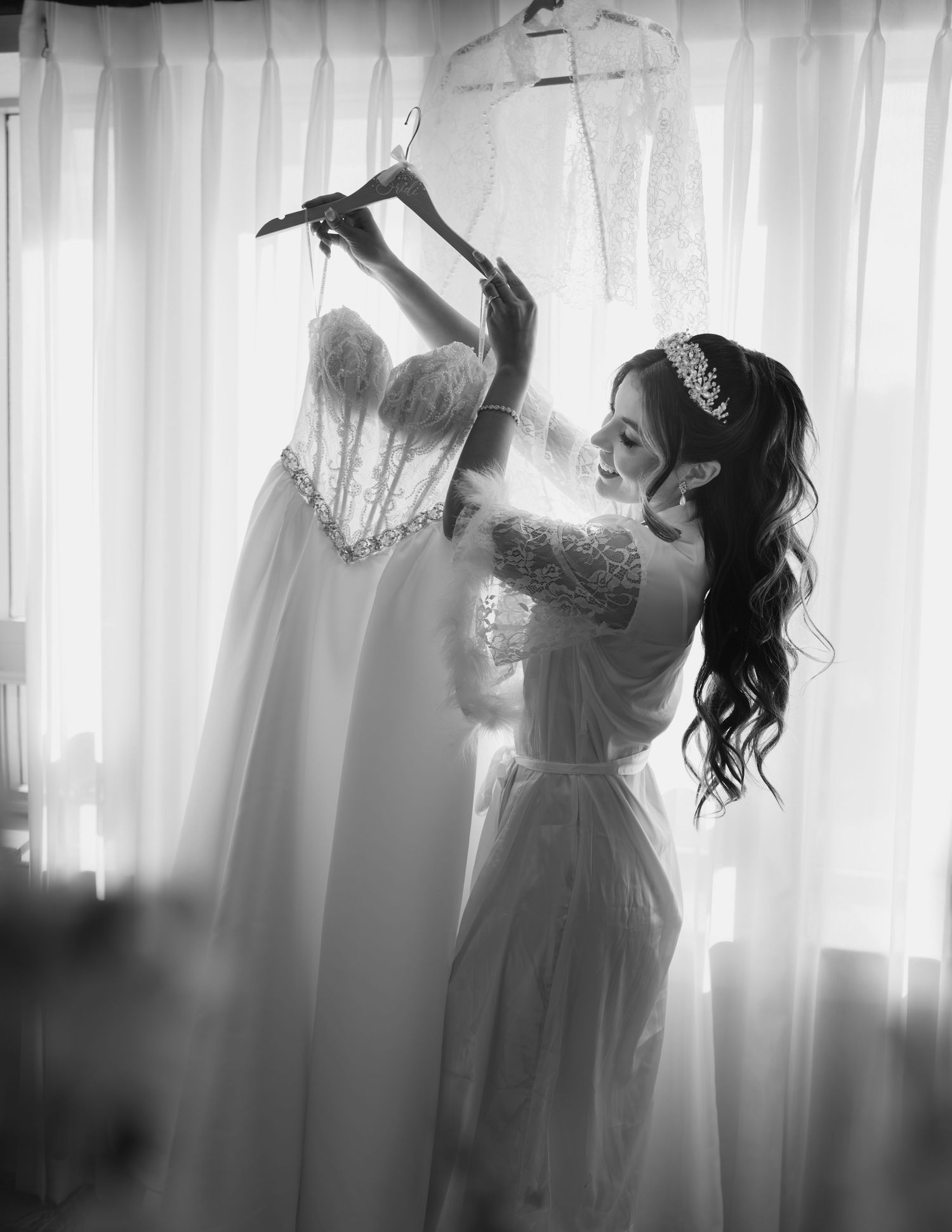 Black and white photo of bride in robe holding pearl wedding dress near sheer curtains.