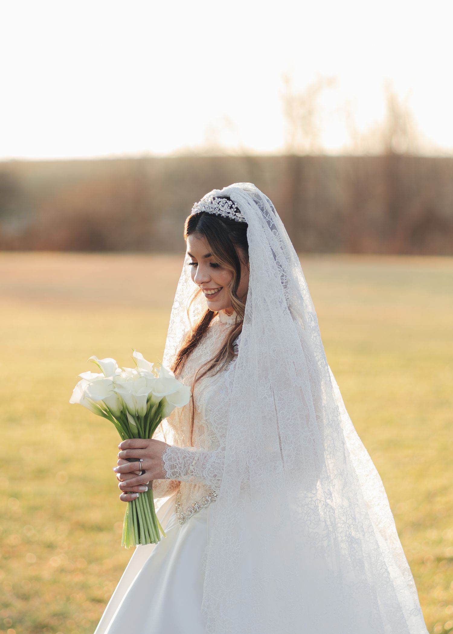 Elegant bride in lace wedding gown and tiara veil smiling while holding white calla lily bouquet.