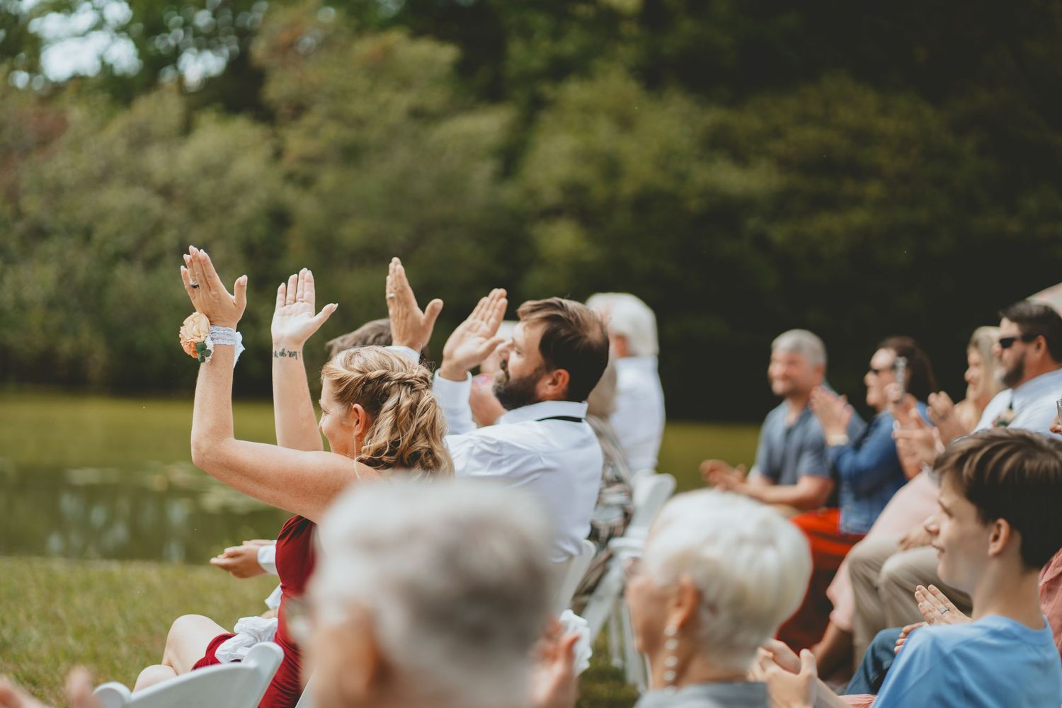 Joyful wedding guests clap and raise hands in celebration at outdoor ceremony by a lake.