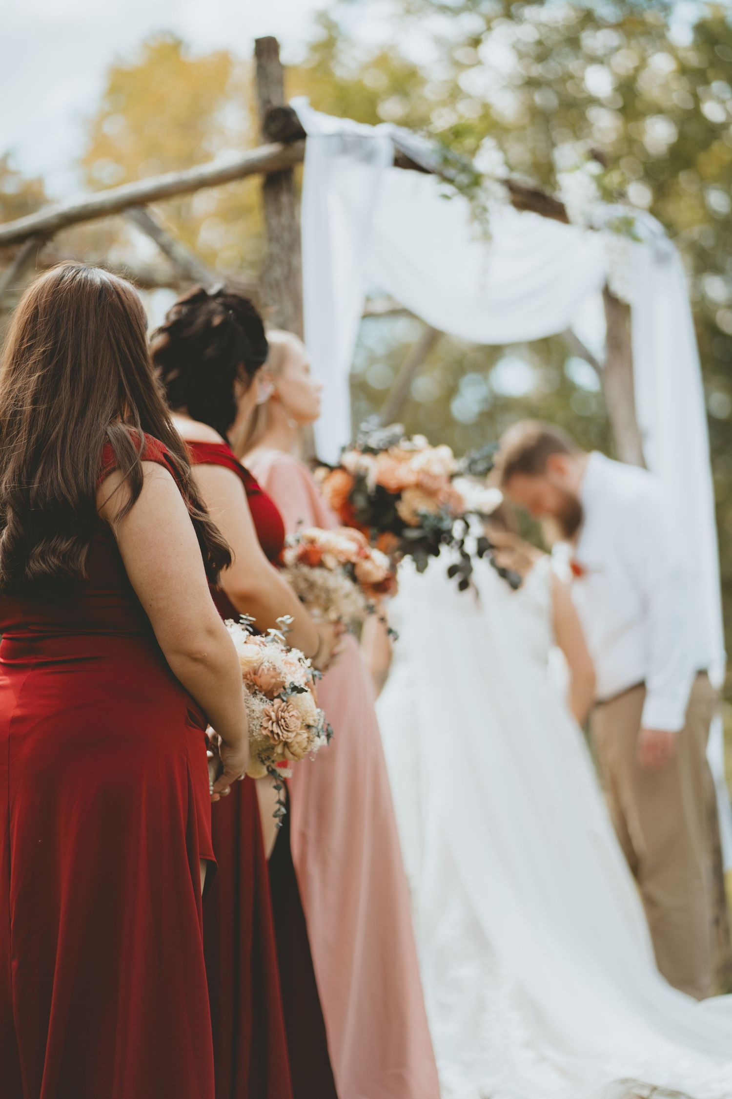 Bridesmaids in red and pink gowns hold bouquets as couple kisses under rustic wooden arch.