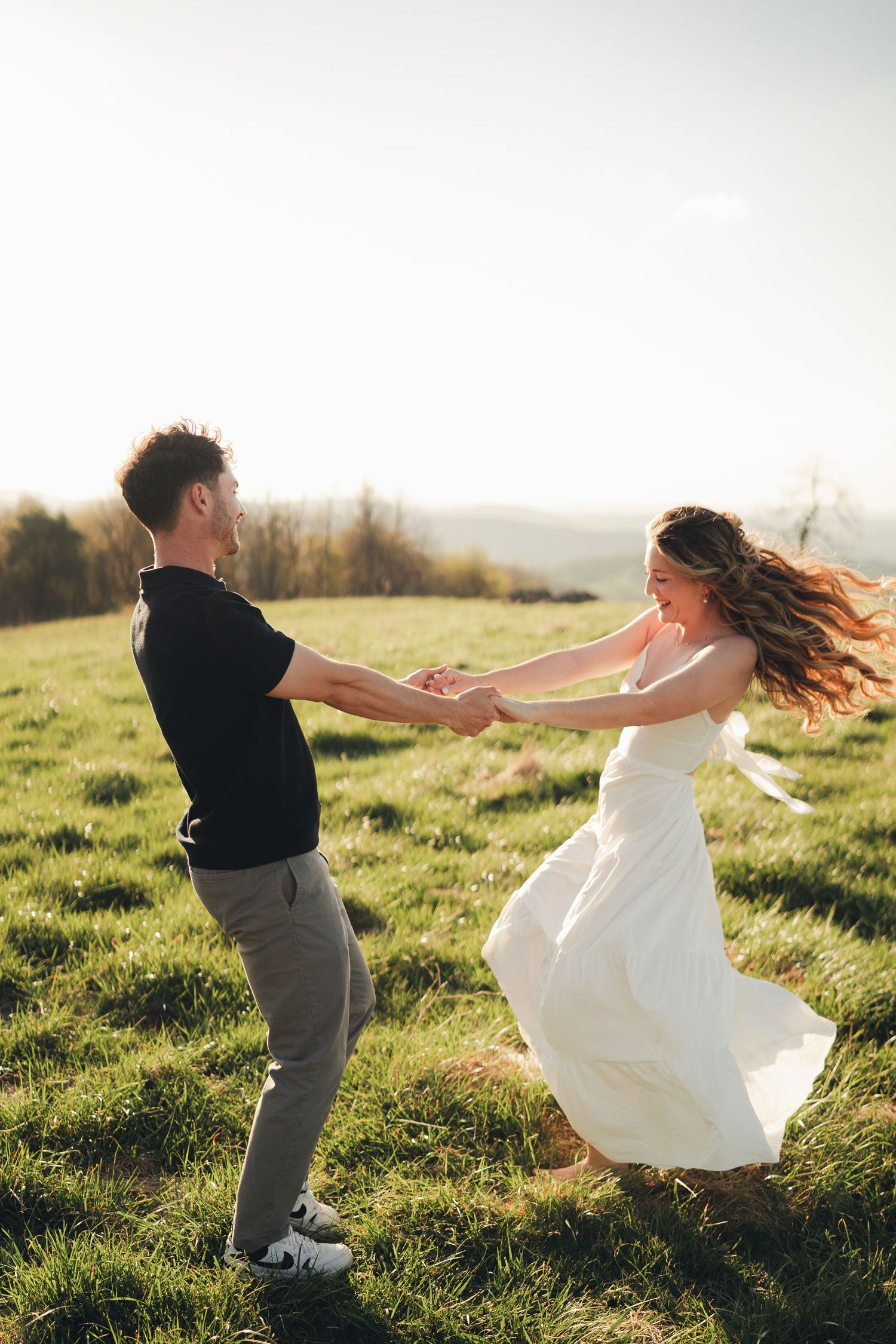 Joyful couple spinning together holding hands on a green hill at sunset.