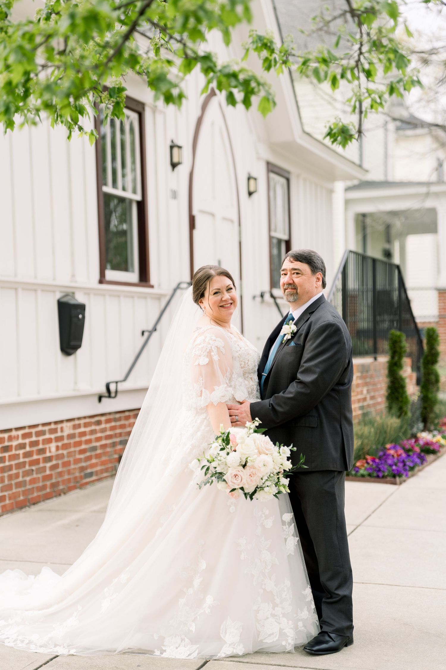 Bride in lace gown and groom in black tux holding hands outside a white wedding venue.