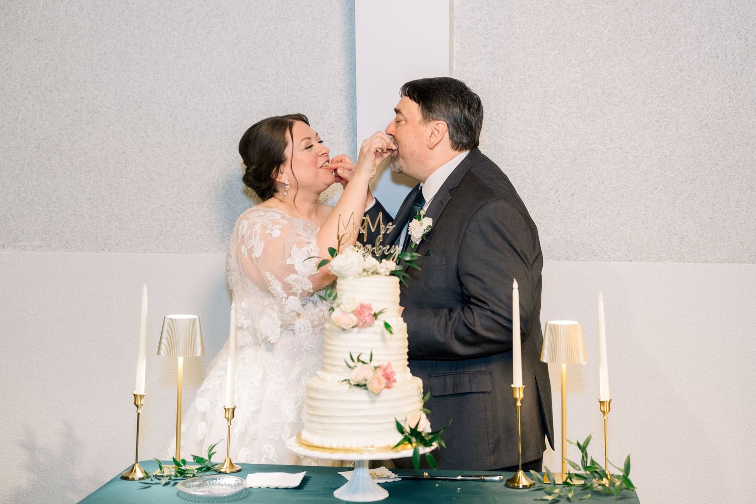 Bride and groom feeding each other wedding cake at elegant reception with champagne flutes and candles.