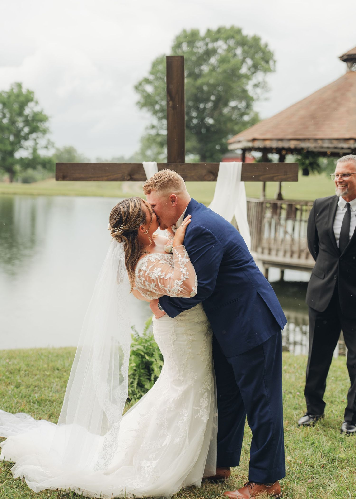 Bride and groom share first kiss at outdoor wedding ceremony by a pond with a wooden cross backdrop.