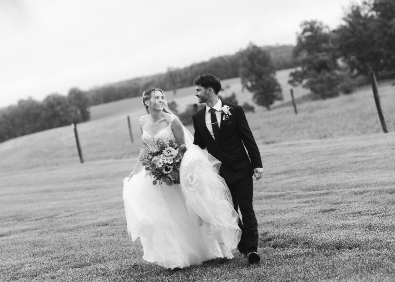 Black and white photo of a bride and groom walking hand in hand across a lush green field on their wedding day.