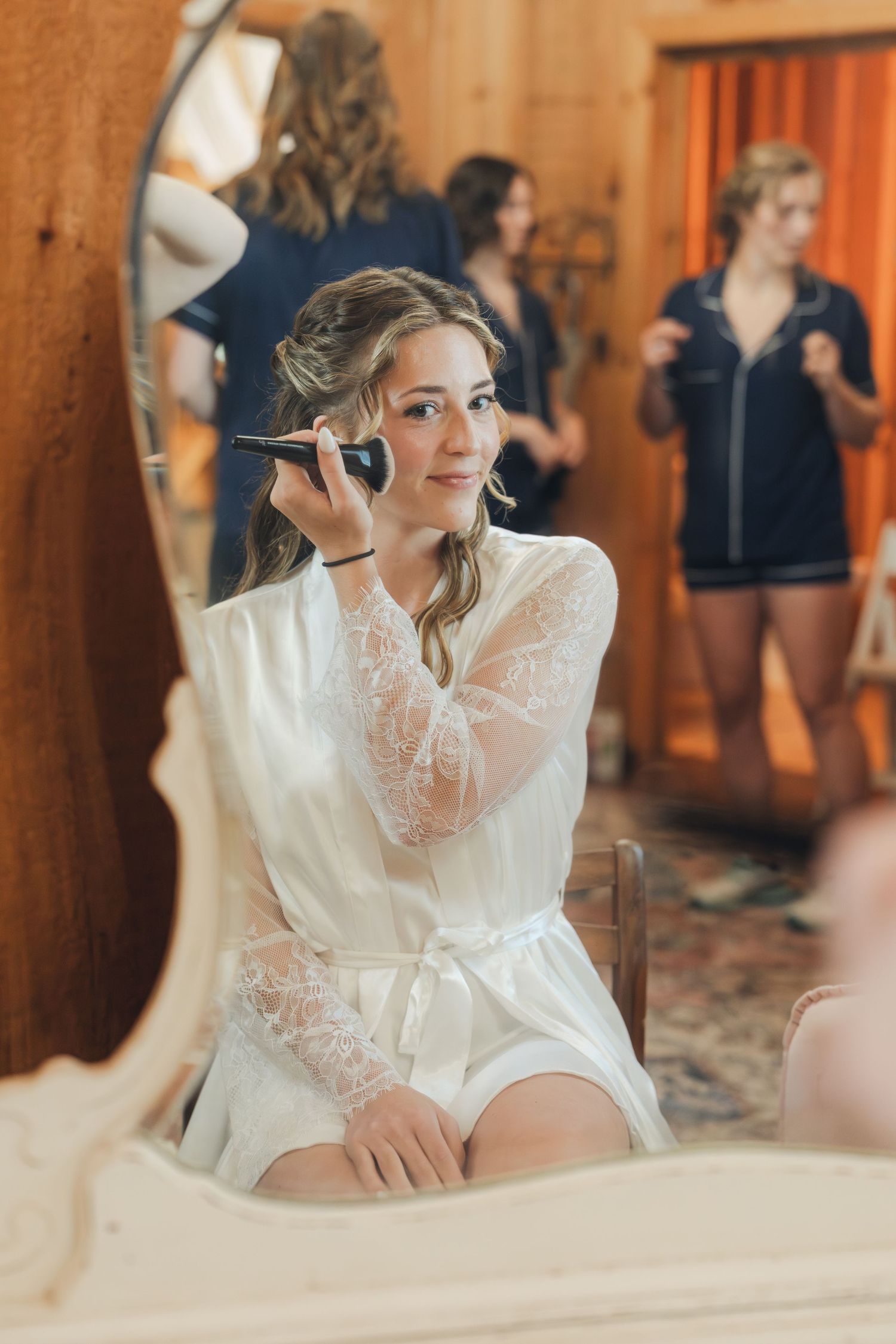 Bride in white lace robe applying makeup at vintage mirror while bridesmaids prepare in background.
