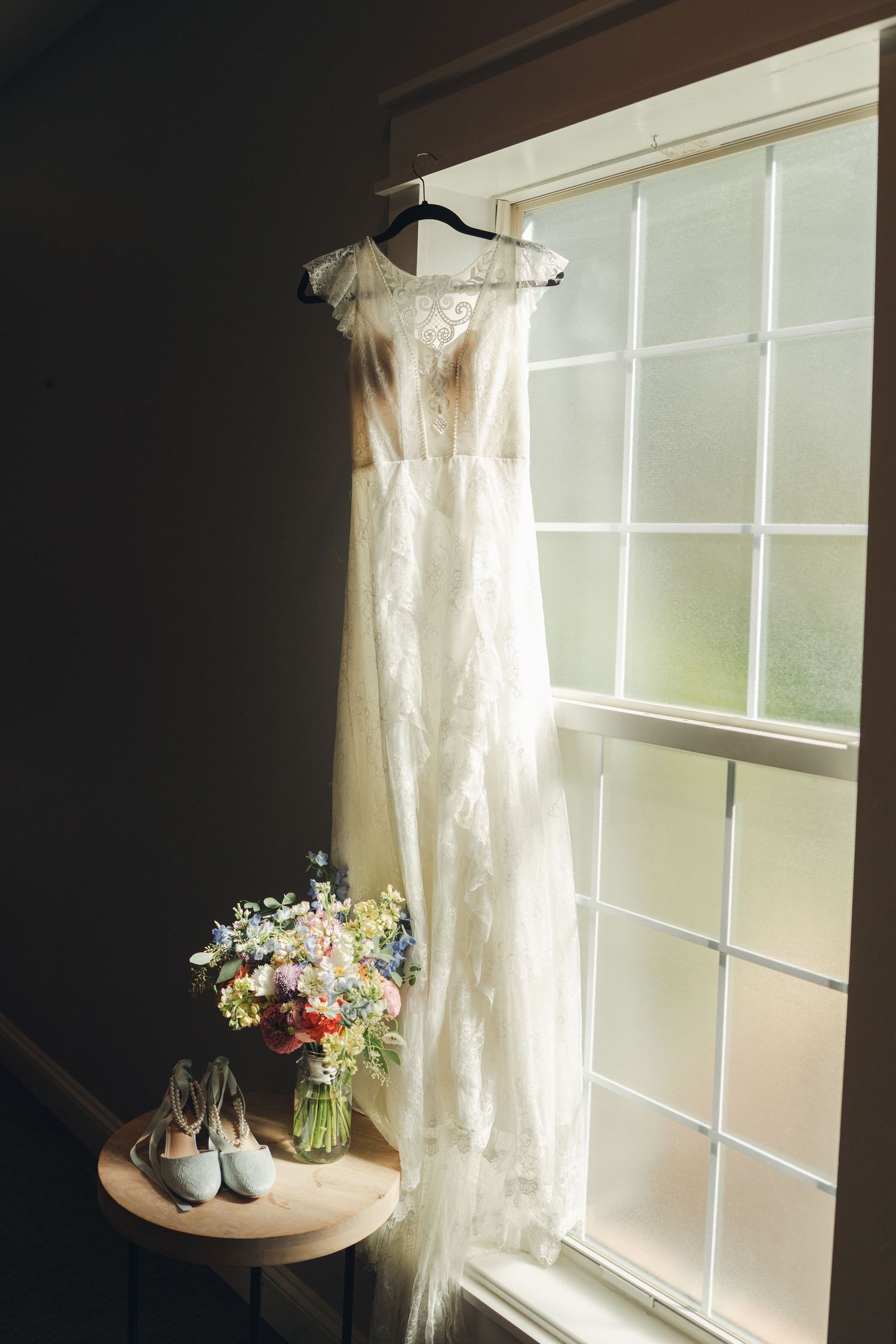 Elegant lace wedding dress hanging by a window with bridal bouquet and blue heels on a wooden table.