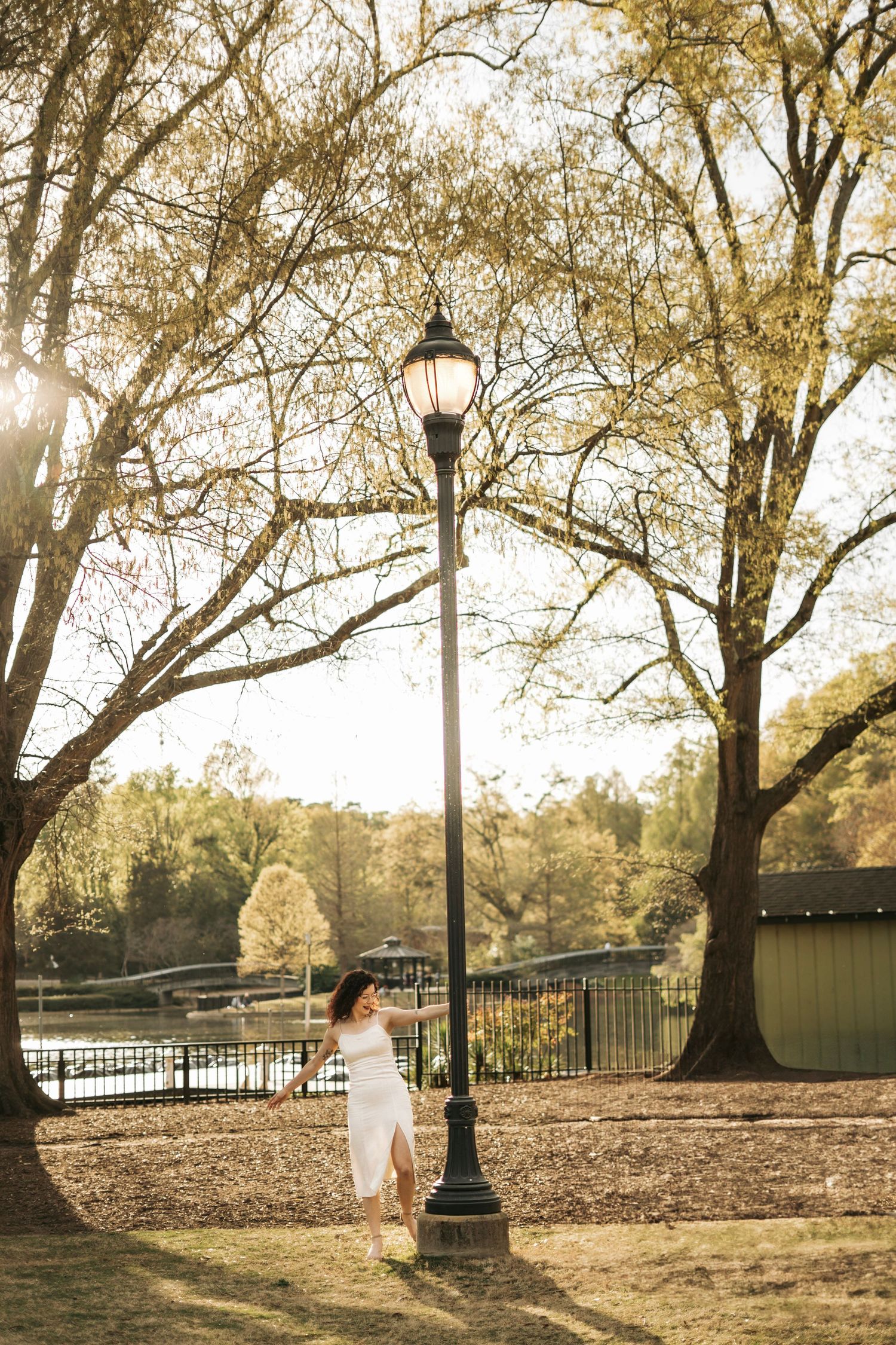 Woman in white dress walking near vintage lamp post in sunlit park with lake backdrop.