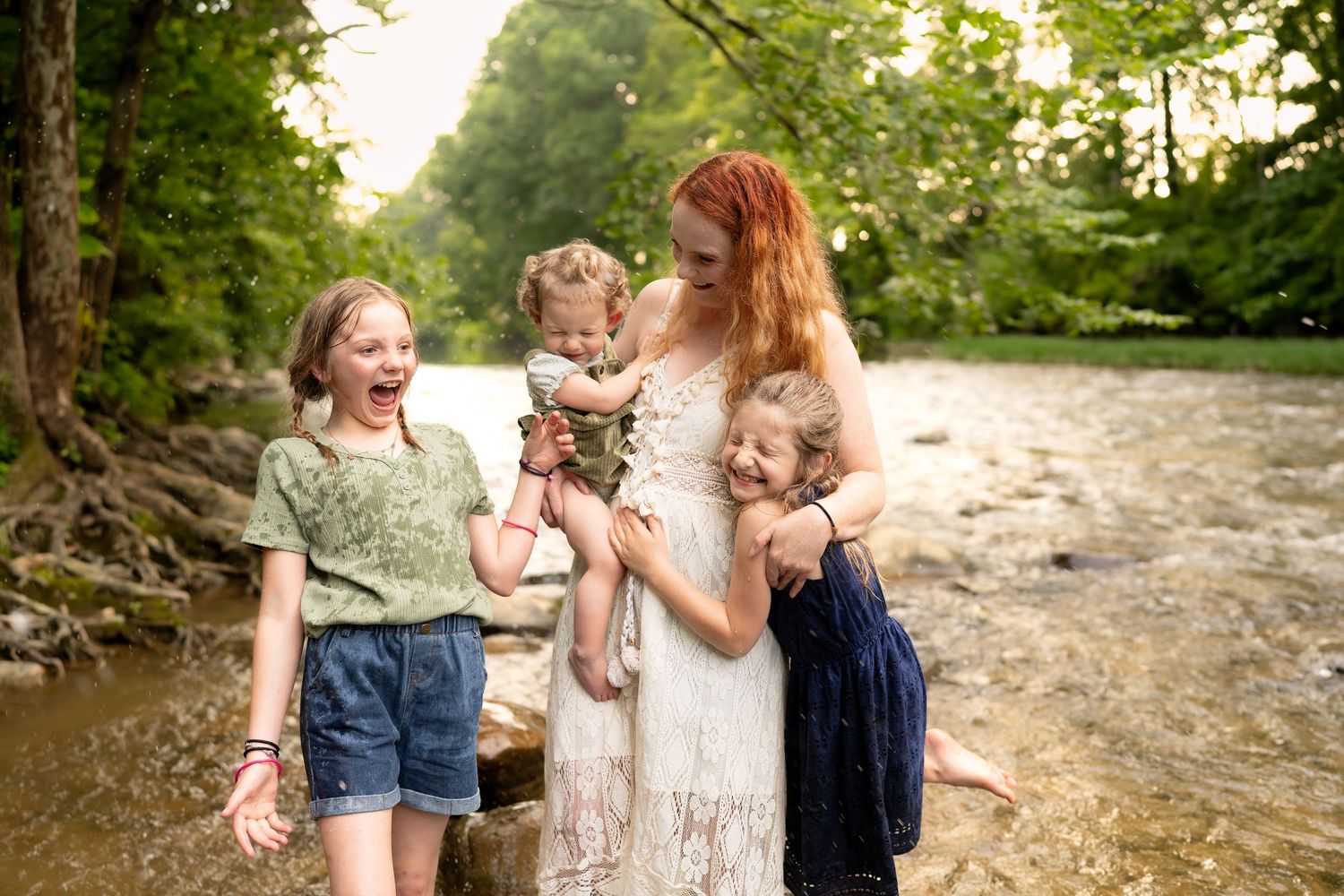 Red-haired mother holding toddler surrounded by three daughters laughing by a scenic river.