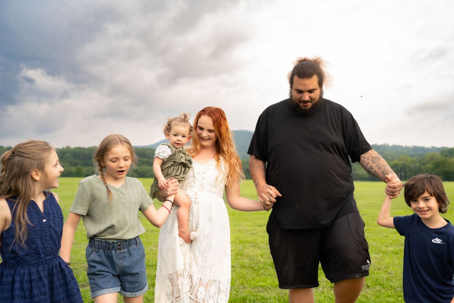 Joyful family of six laughing together outdoors in a green meadow with rolling hills in background.