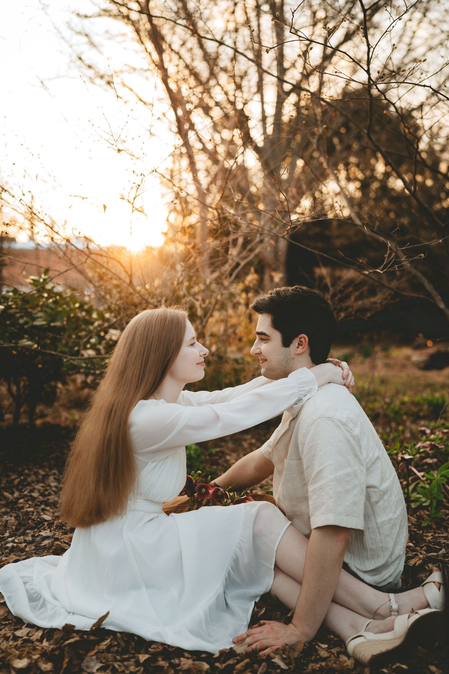 Couple sitting on autumn leaves at golden hour, woman in white dress tenderly touching mans shoulders.
