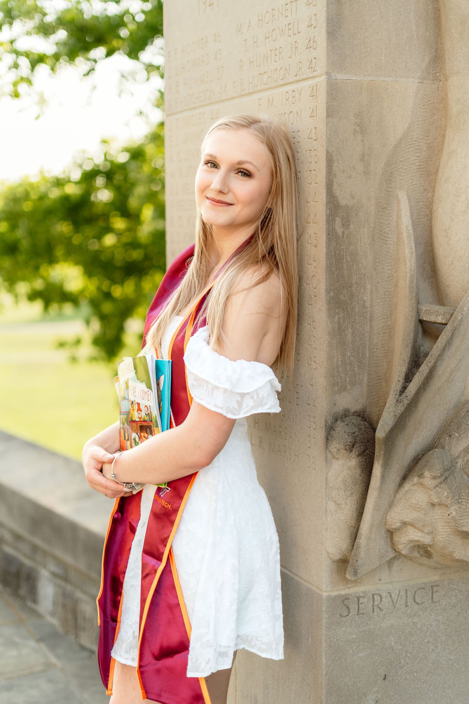 Blonde graduate in white dress and red stole leans against a stone column engraved with SERVICE.